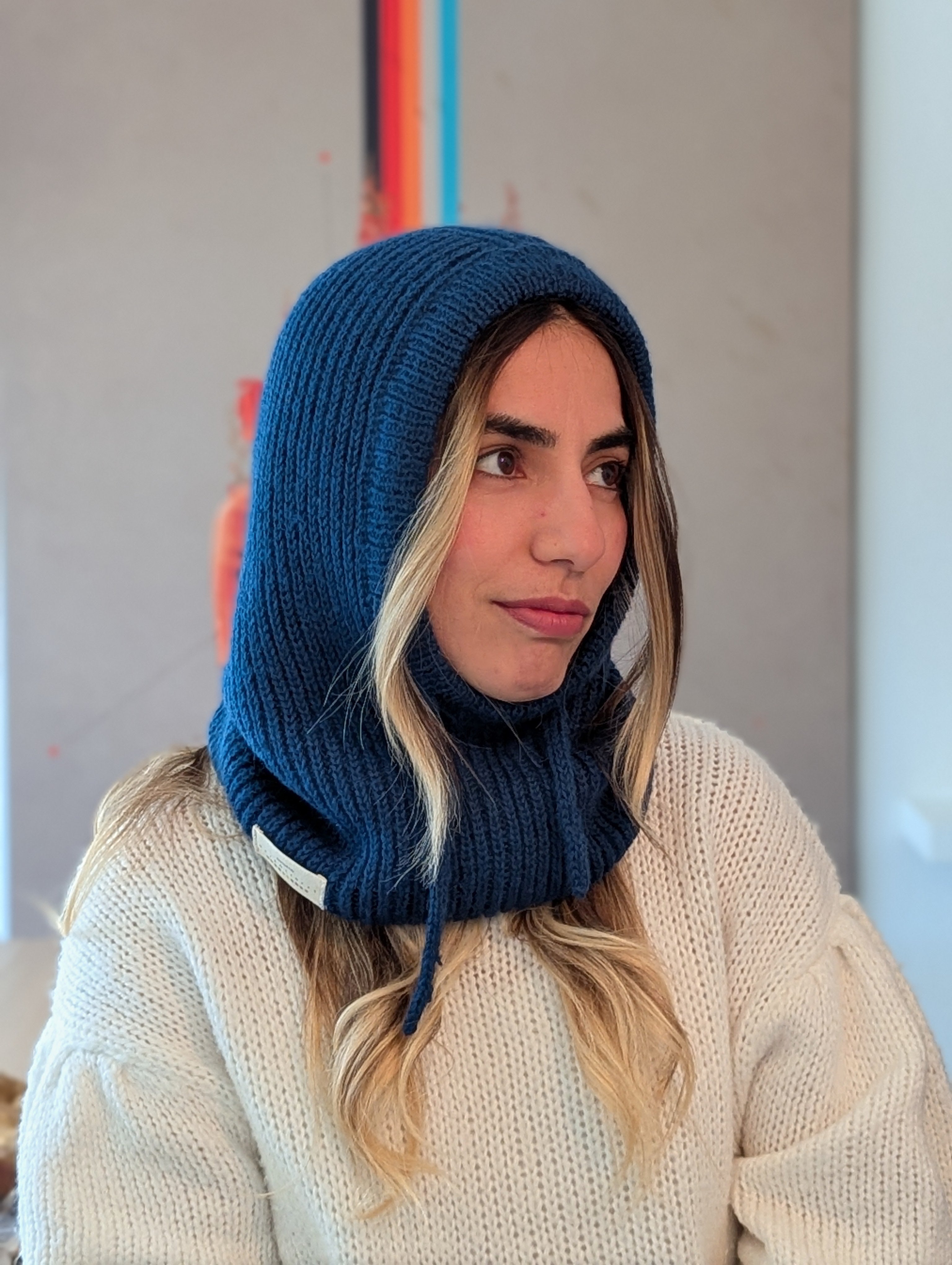 Woman wearing a blue knitted hood over her head, sitting indoors.