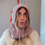 Person wearing a pink knitted hood and scarf blowing a kiss against a neutral background
