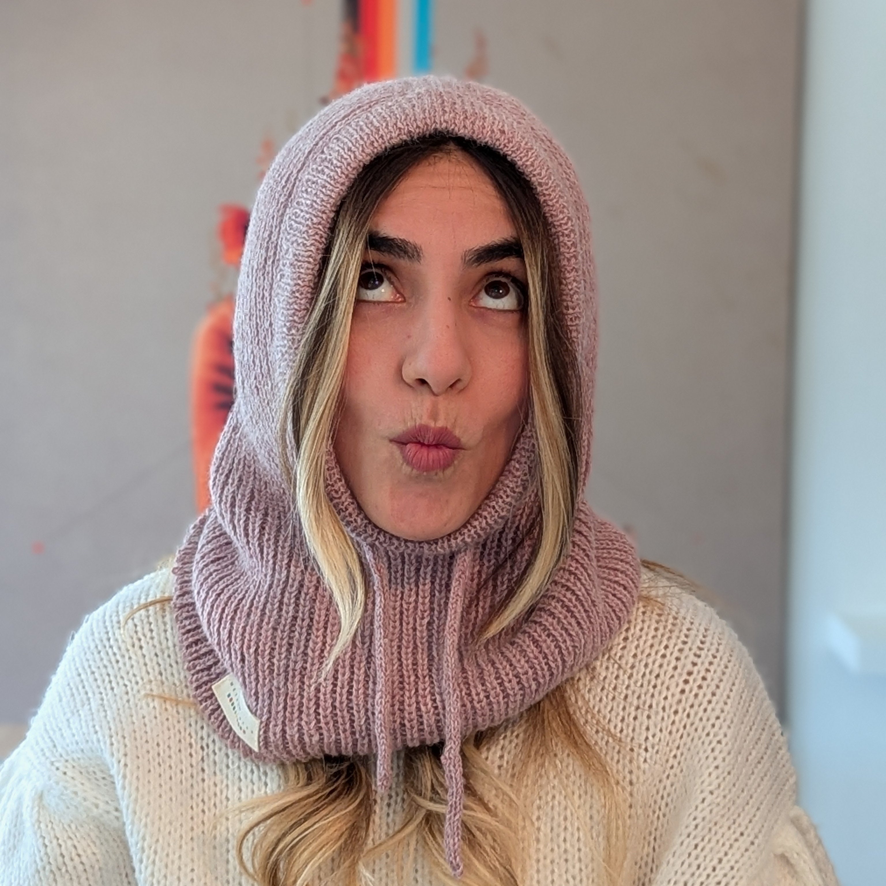 Person wearing a pink knitted hood and scarf blowing a kiss against a neutral background