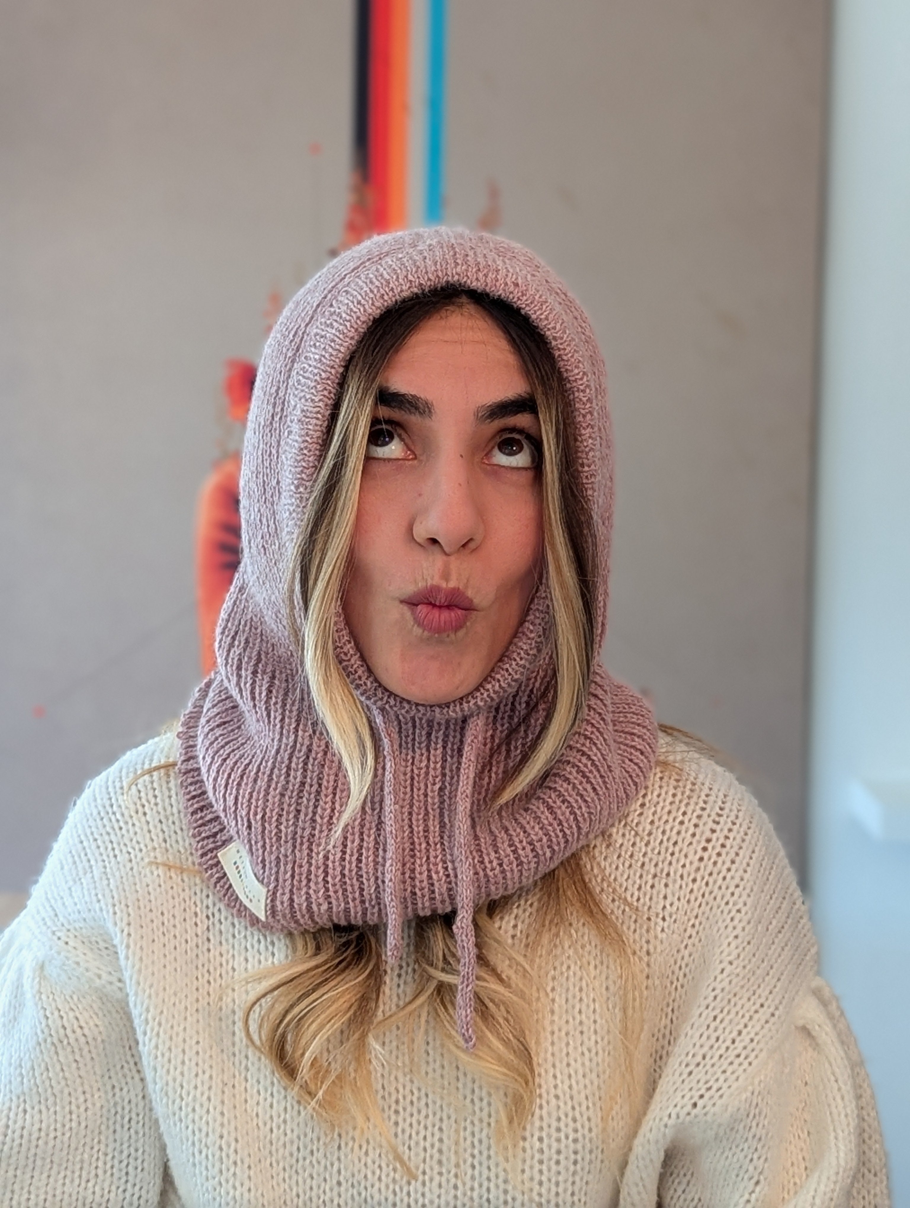 Person wearing a pink knitted hood and scarf blowing a kiss against a neutral background