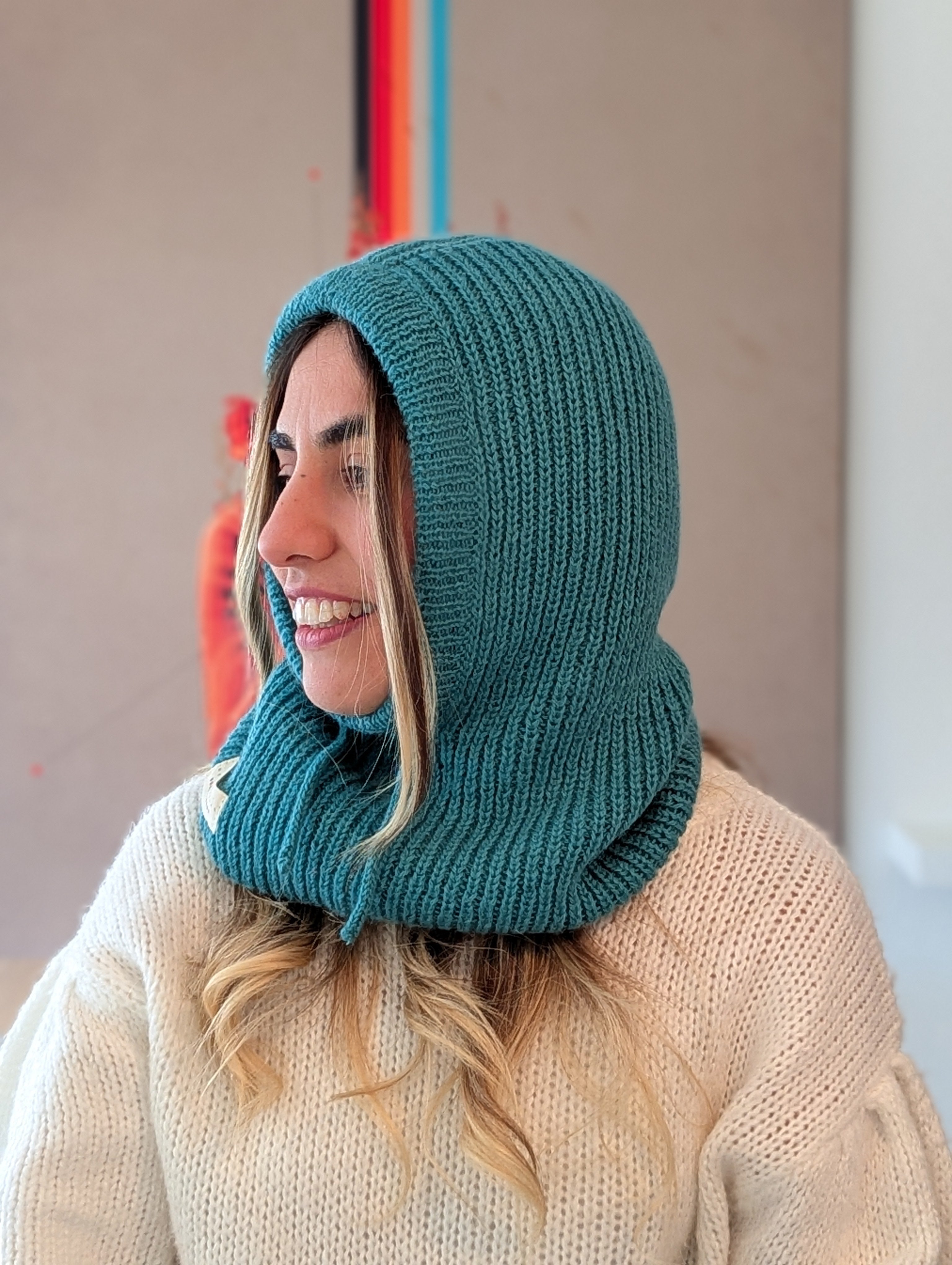 Woman wearing a teal knitted hood over her head with a beige sweater underneath.