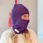 Person wearing a purple knitted balaclava with a brand label, standing against a neutral background.