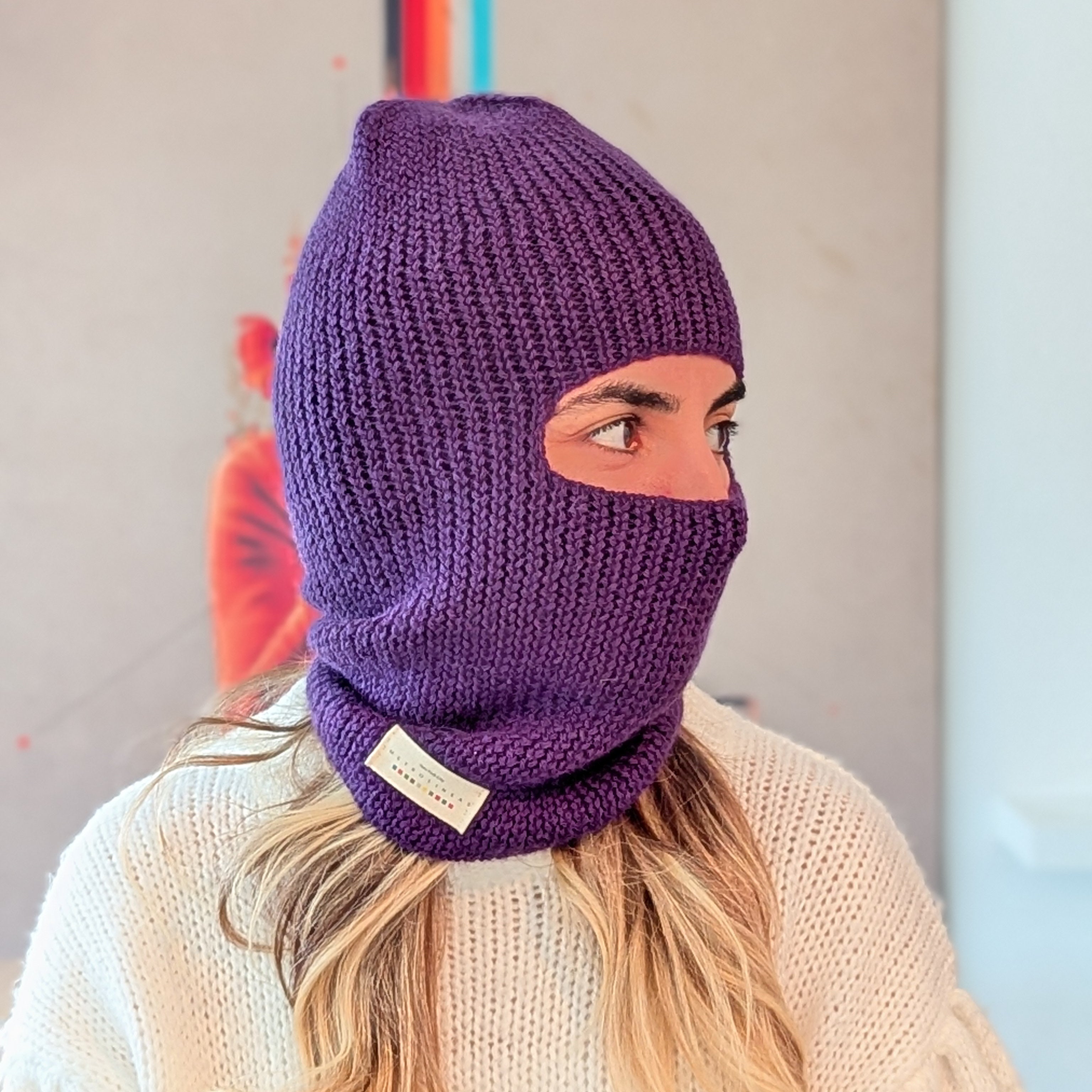 Person wearing a purple knitted balaclava with a brand label, standing against a neutral background.
