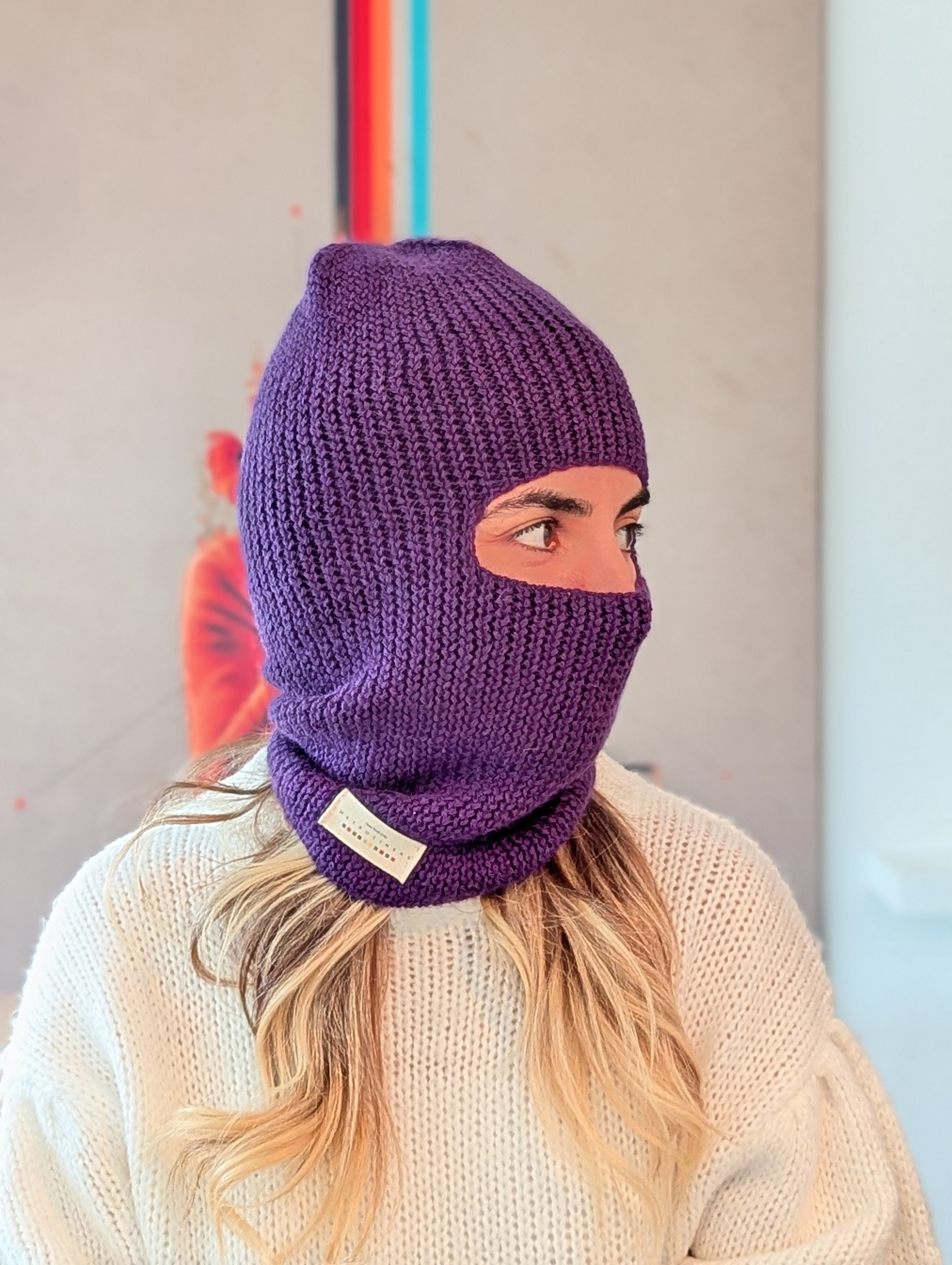 Person wearing a purple knitted balaclava with a brand label, standing against a neutral background.