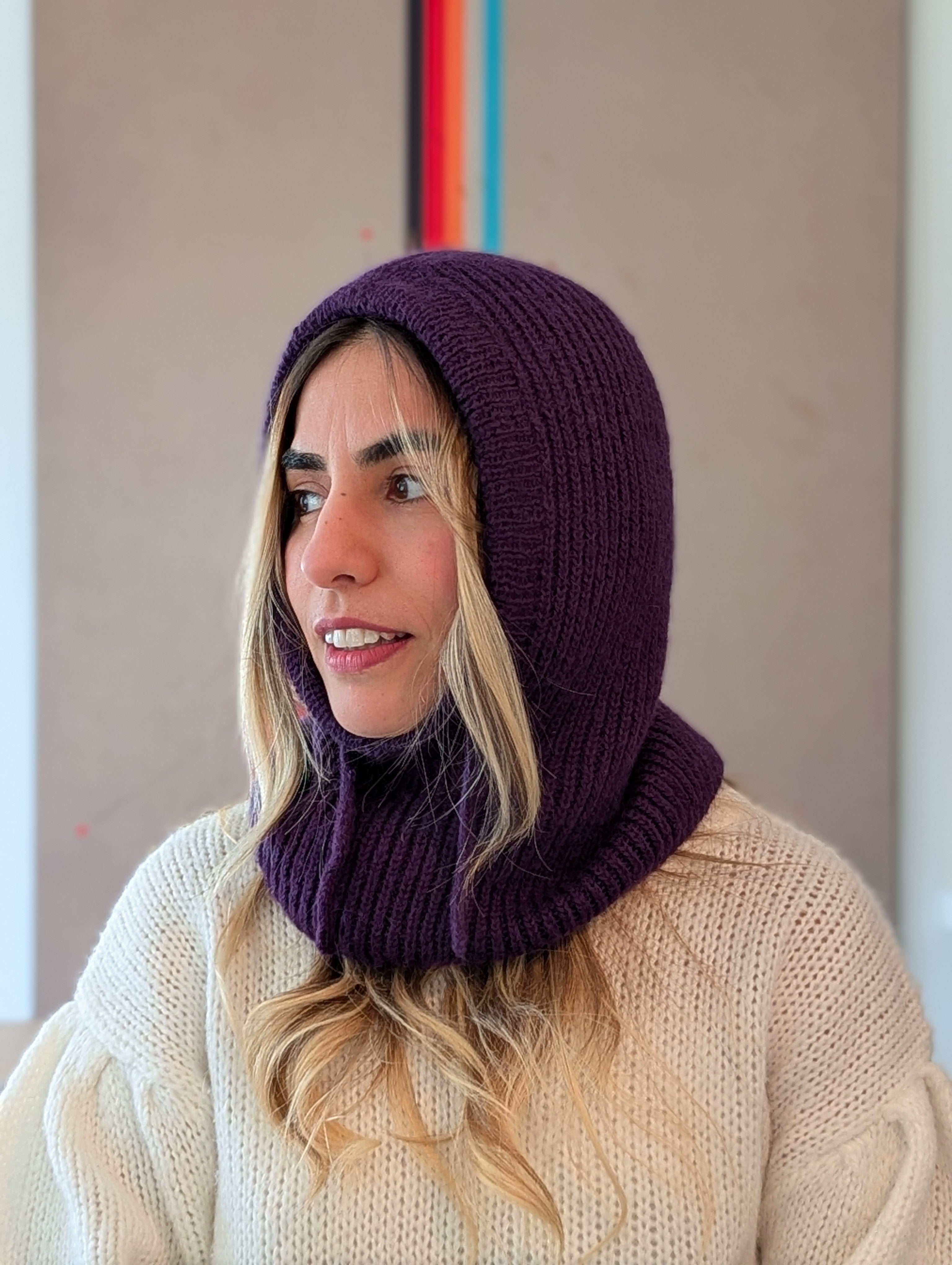 Woman wearing a purple knitted hood and beige sweater against a neutral background