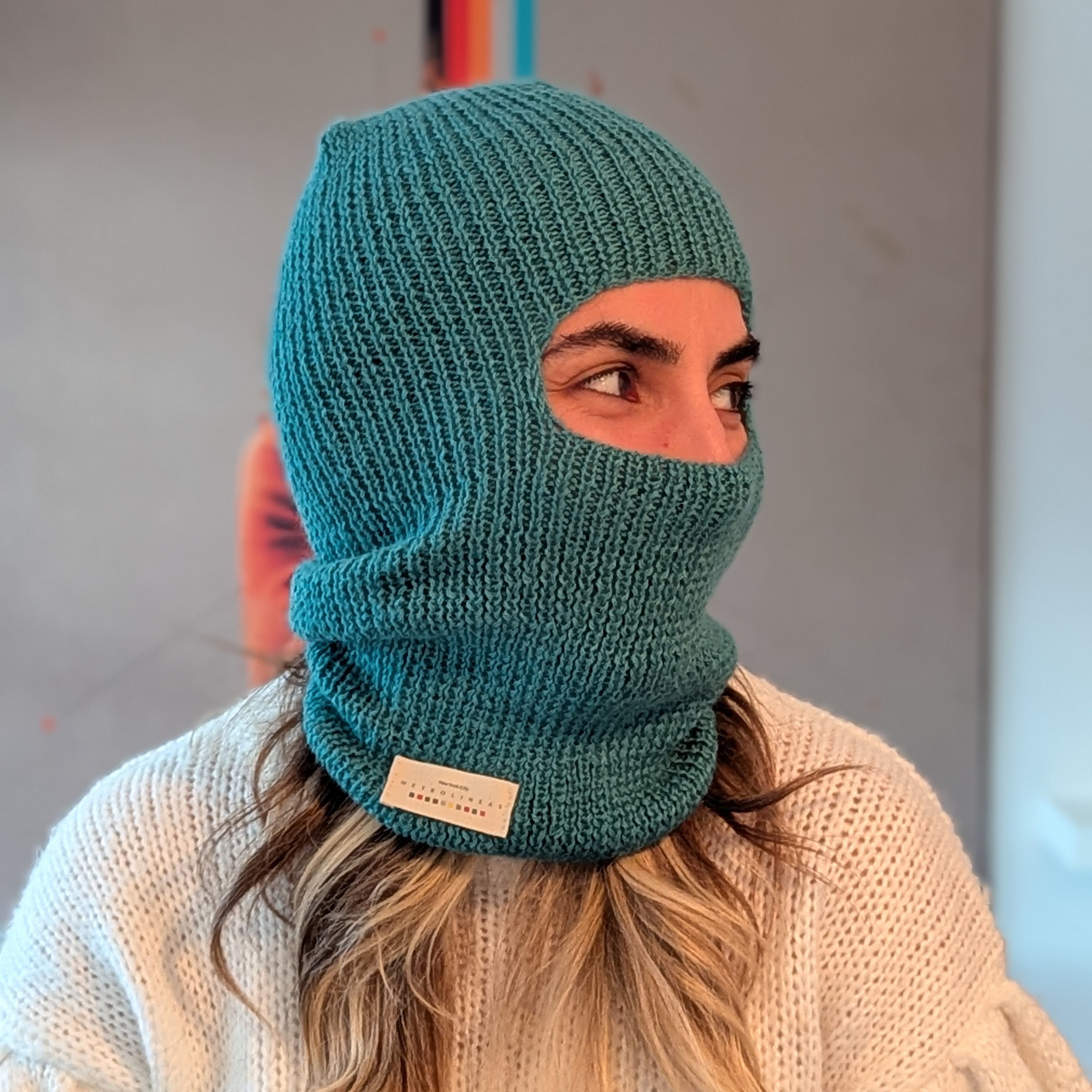 Person wearing a teal knitted balaclava with a visible brand label against a neutral background