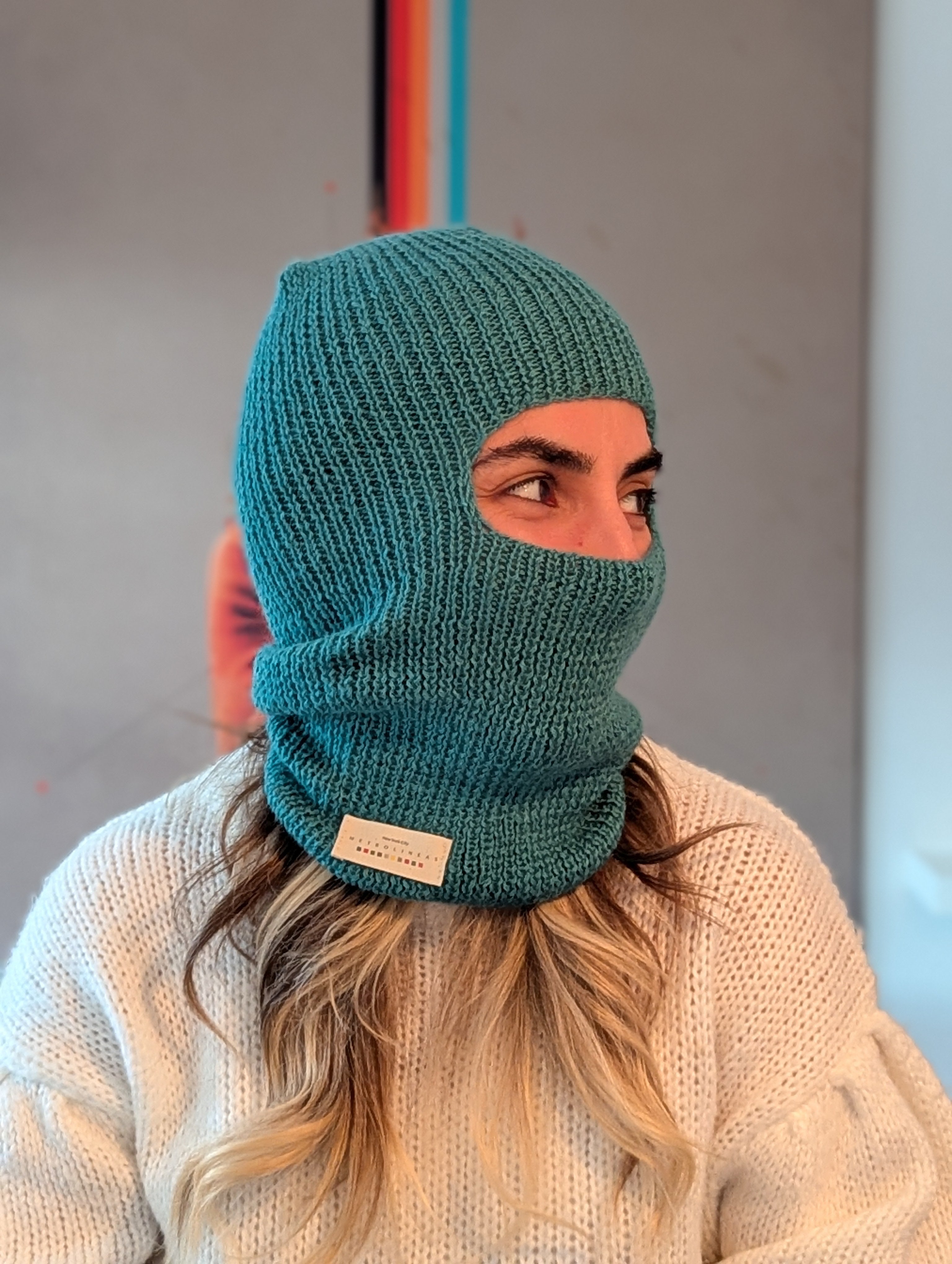 Person wearing a teal knitted balaclava with a visible brand label against a neutral background