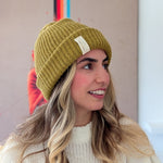 Woman wearing a mustard yellow beanie with a brand logo, smiling against a neutral background.