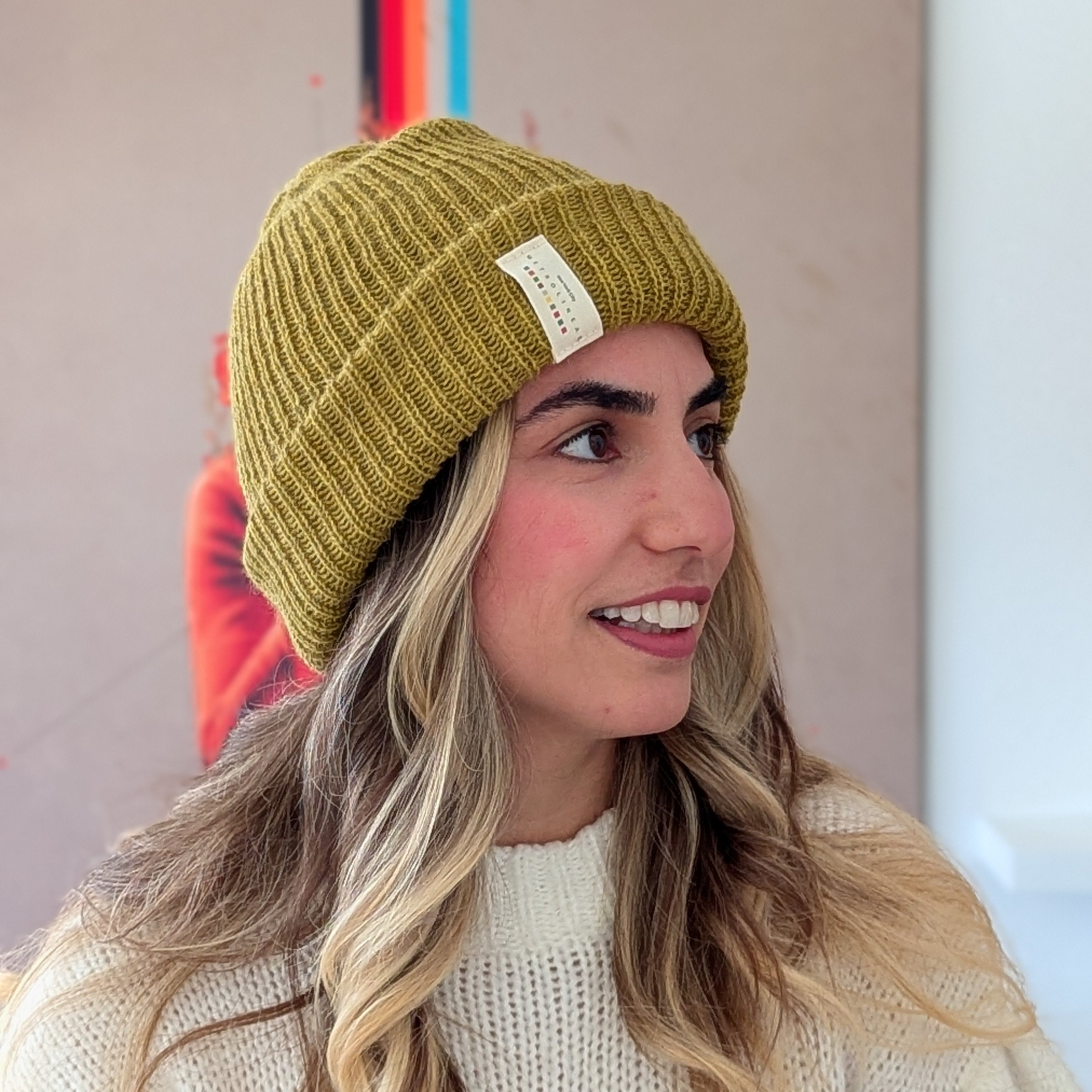 Woman wearing a mustard yellow beanie with a brand logo, smiling against a neutral background.