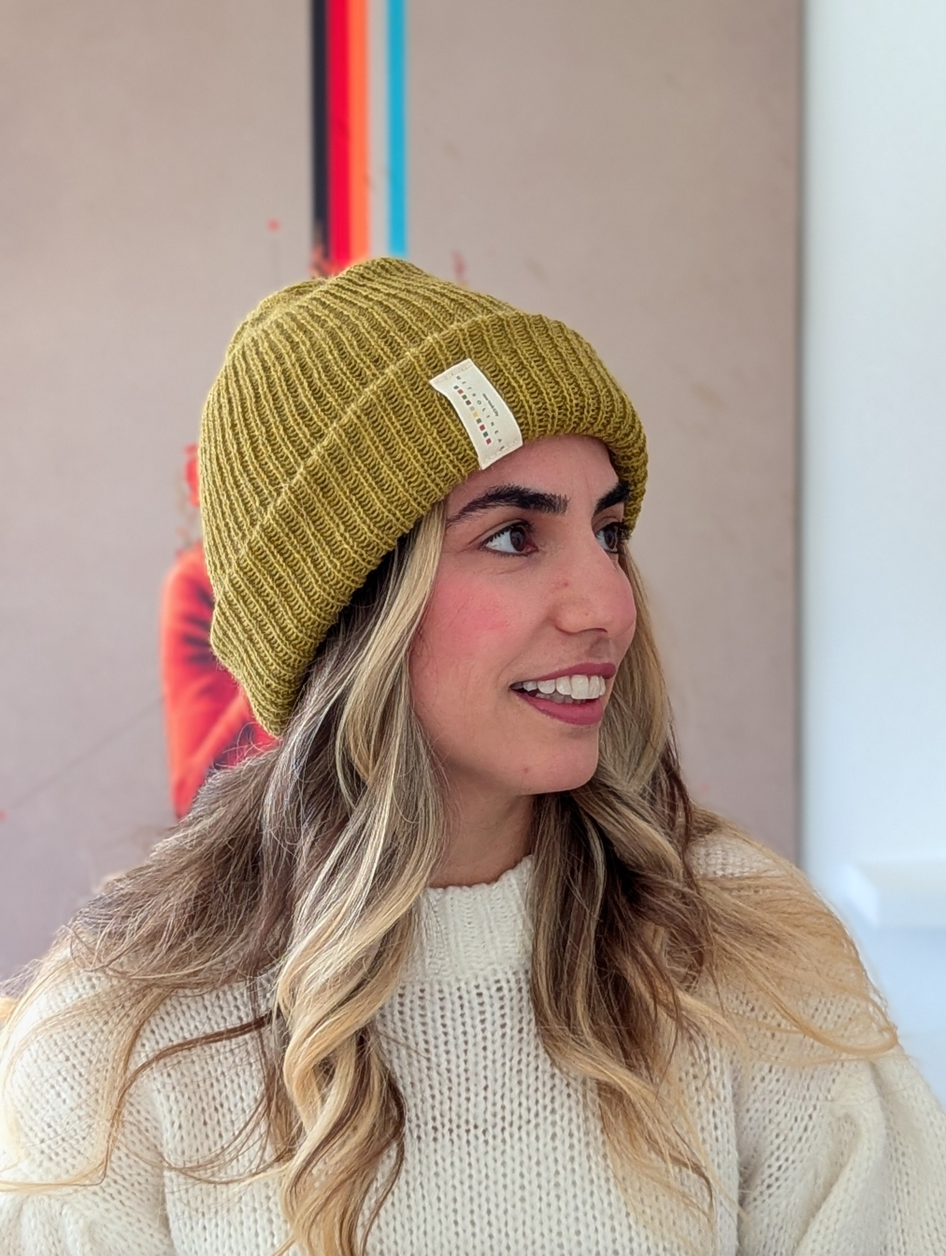 Woman wearing a mustard yellow beanie with a brand logo, smiling against a neutral background.