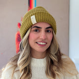 Person wearing a mustard yellow beanie with a brand tag, smiling in front of a colorful abstract painting.