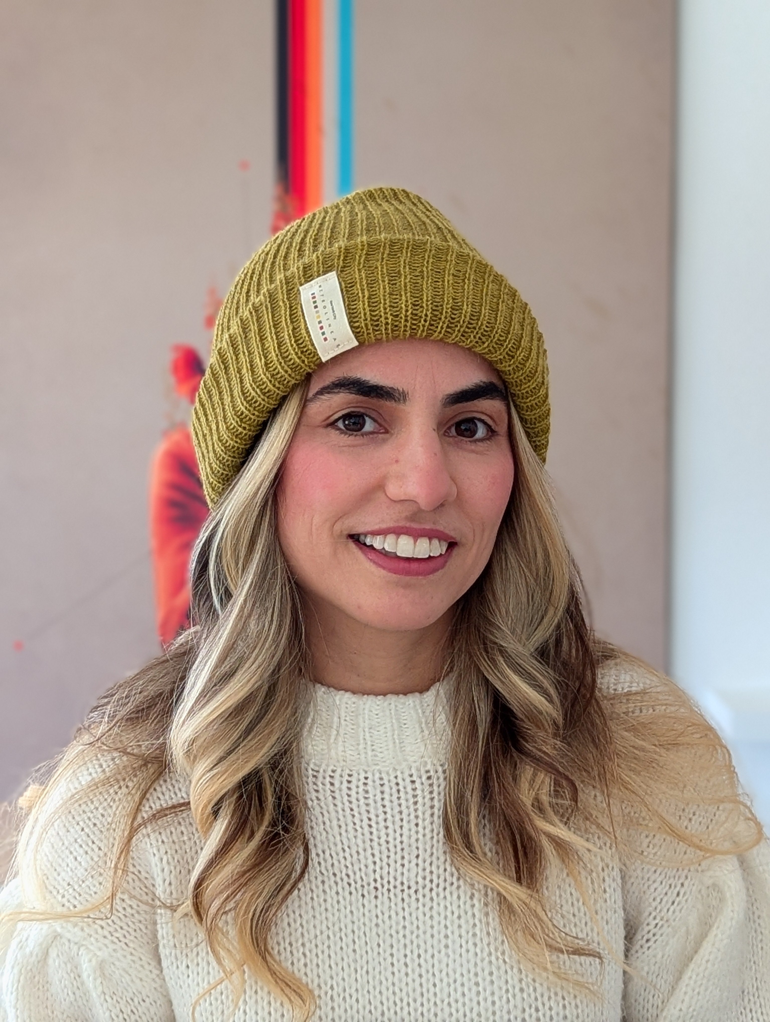 Person wearing a mustard yellow beanie with a brand tag, smiling in front of a colorful abstract painting.