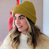 Woman wearing a mustard yellow knit beanie and white sweater against a neutral background