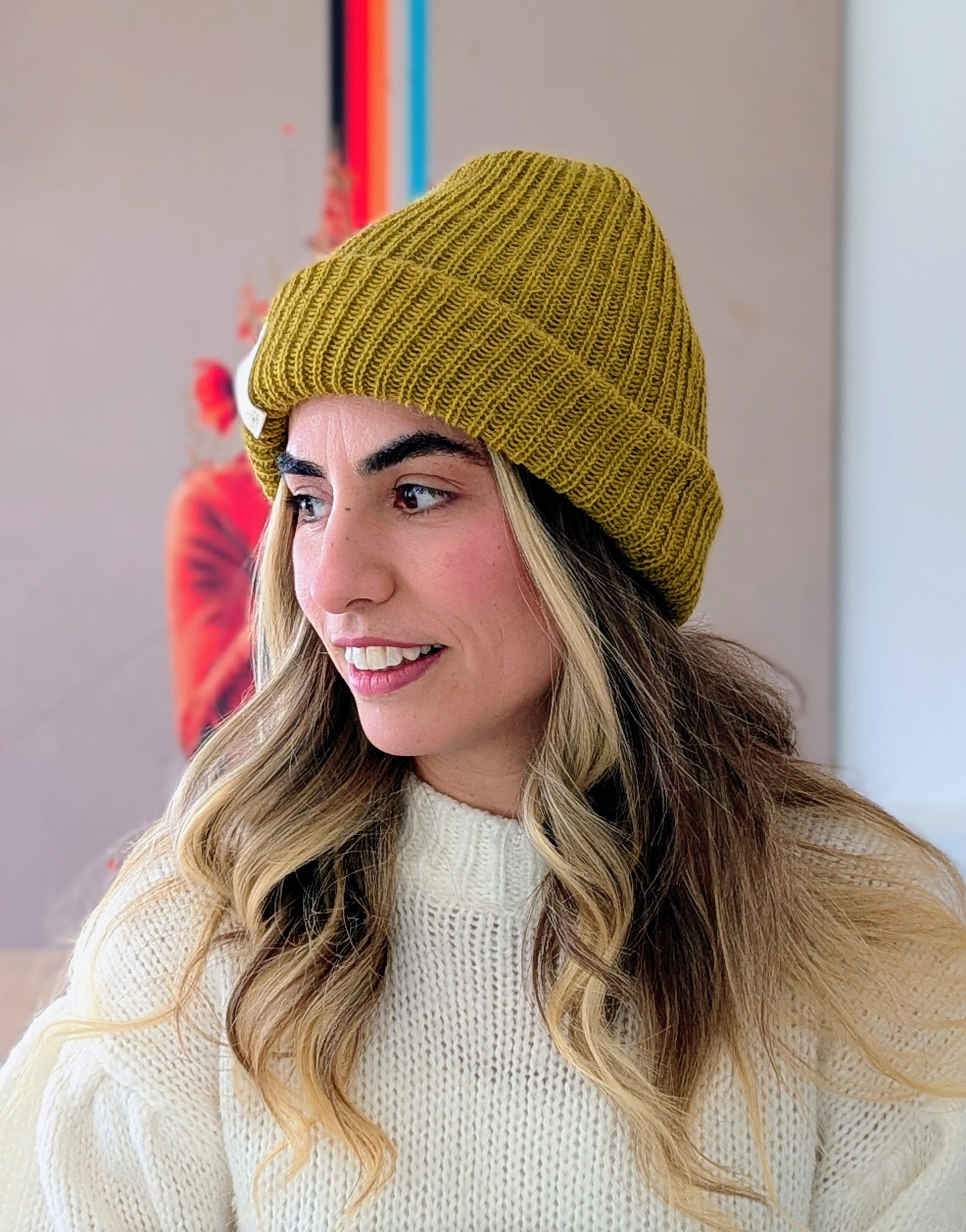 Woman wearing a mustard yellow knit beanie and white sweater against a neutral background