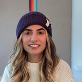 Woman wearing a purple beanie and white sweater with a colorful abstract painting in the background