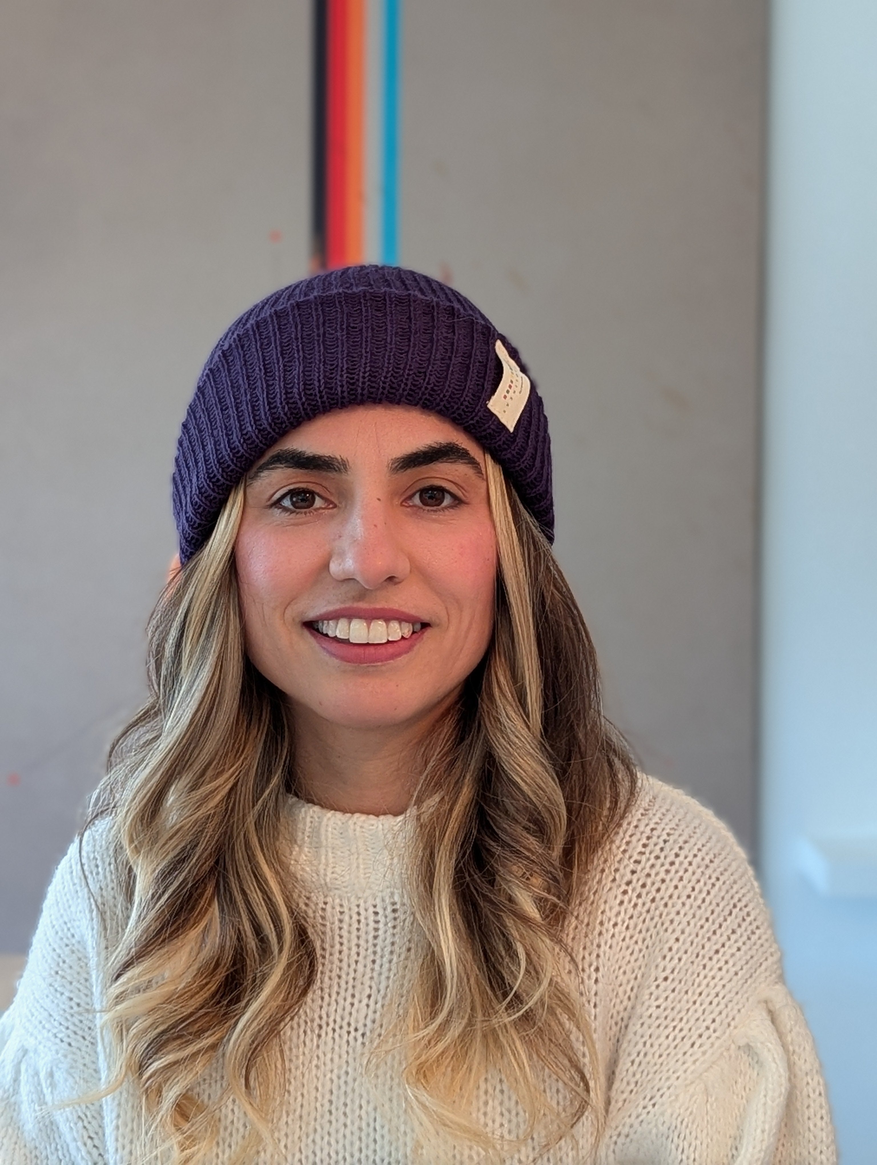Woman wearing a purple beanie and white sweater with a colorful abstract painting in the background