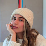 Woman wearing a white knit beanie and sweater indoors with a colorful abstract painting in the background.