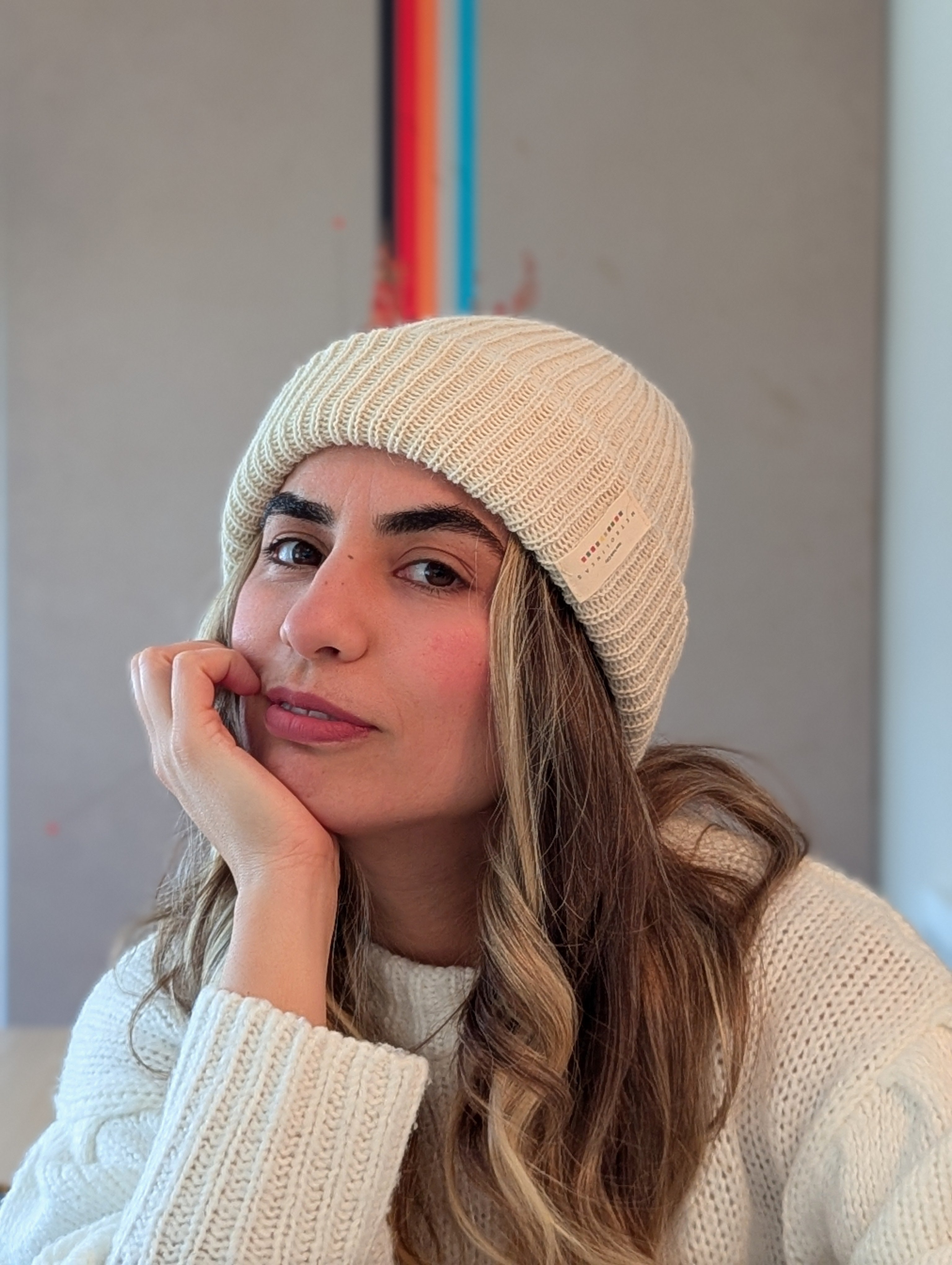 Woman wearing a white knit beanie and sweater indoors with a colorful abstract painting in the background.