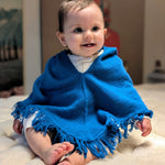 Baby wearing a blue poncho sitting on a light-colored surface.