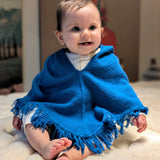 Baby wearing a blue poncho sitting on a light-colored surface.