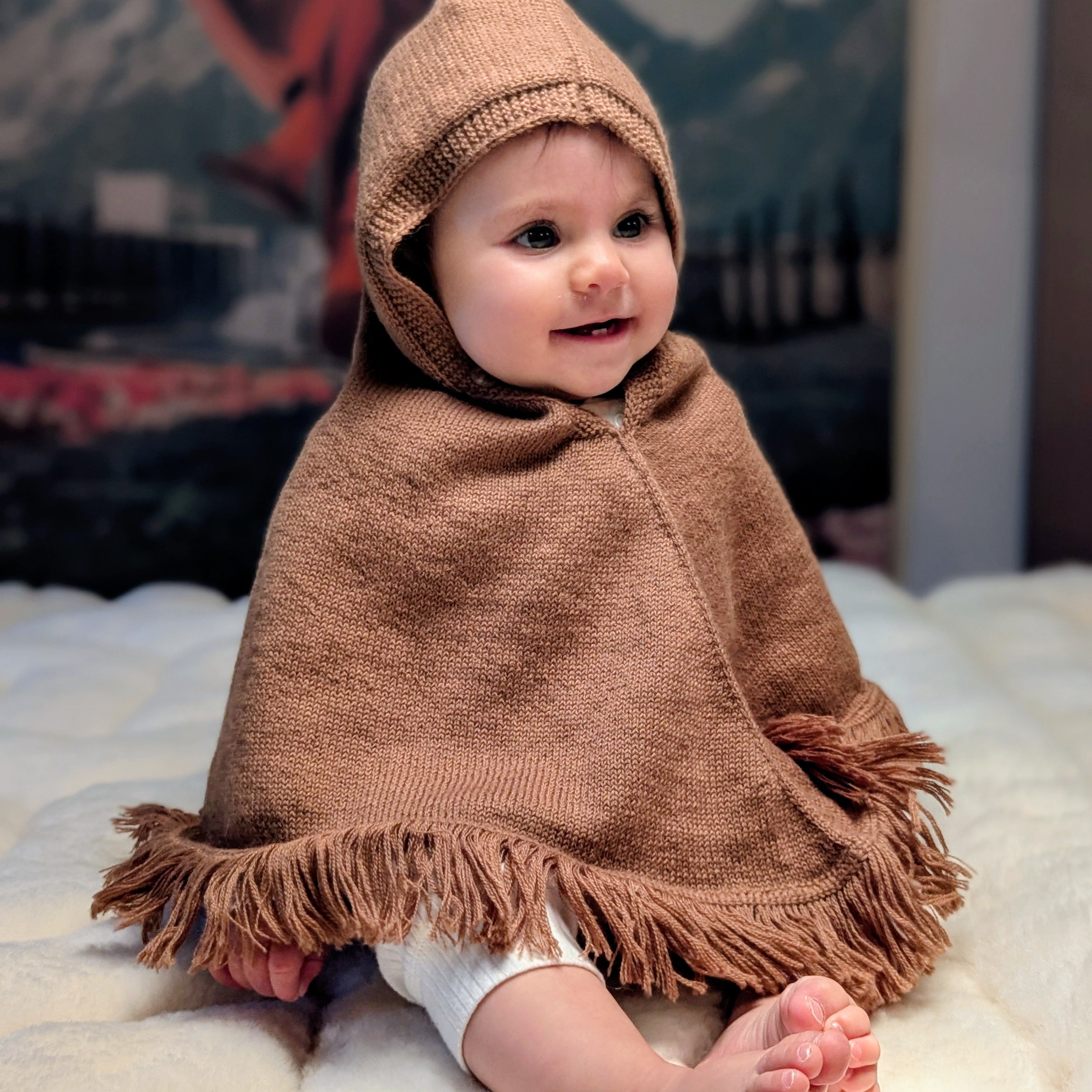 Baby wearing a brown hooded poncho sitting on a white surface with a mountainous background.