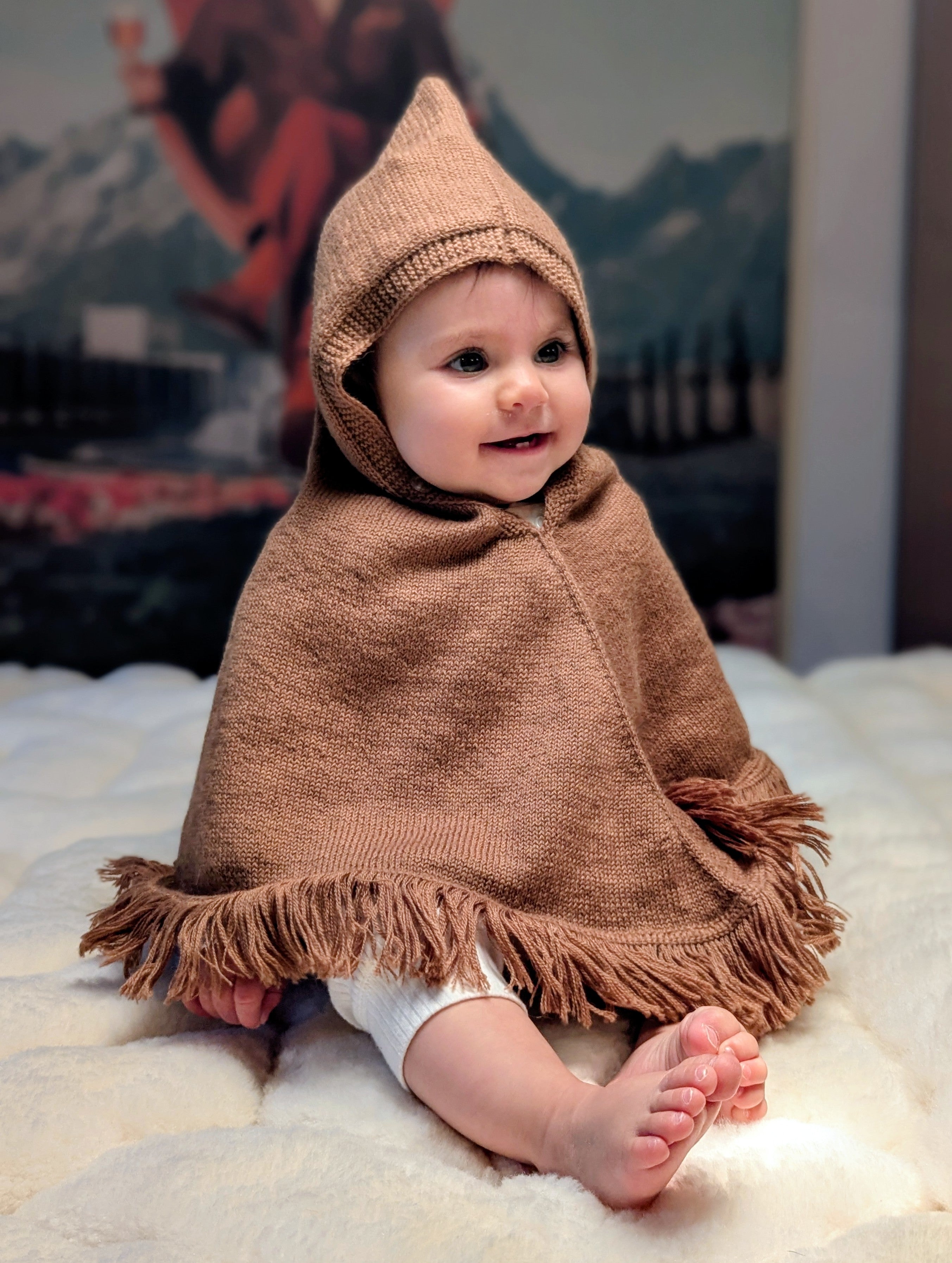 Baby wearing a brown hooded poncho sitting on a white surface with a mountainous background.