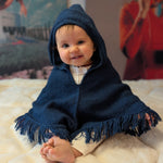 Baby wearing a blue hooded poncho sitting on a white surface with colorful abstract art in the background.