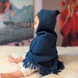 Child wearing a blue hooded poncho sitting on a soft surface with a colorful abstract painting in the background.