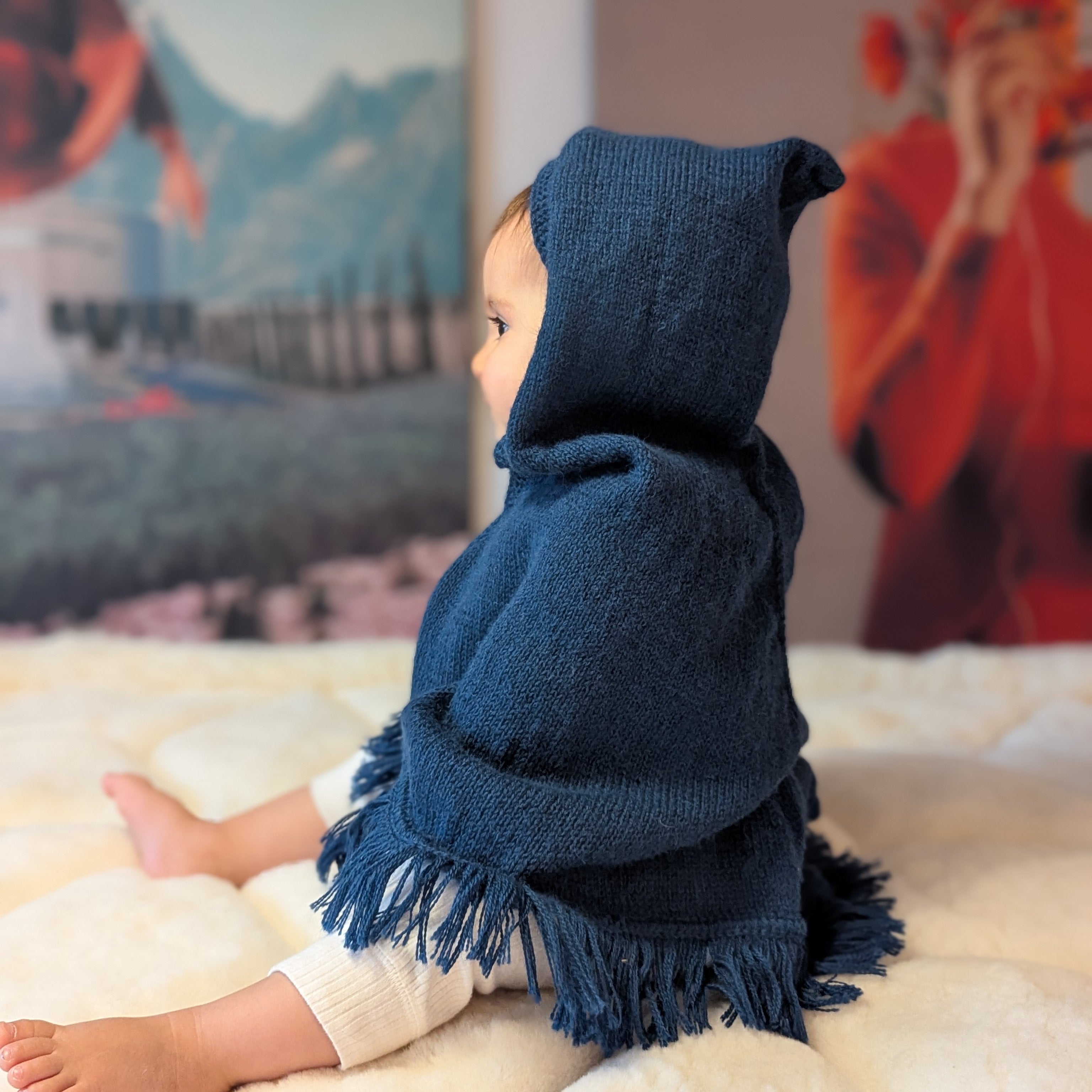 Child wearing a blue hooded poncho sitting on a soft surface with a colorful abstract painting in the background.