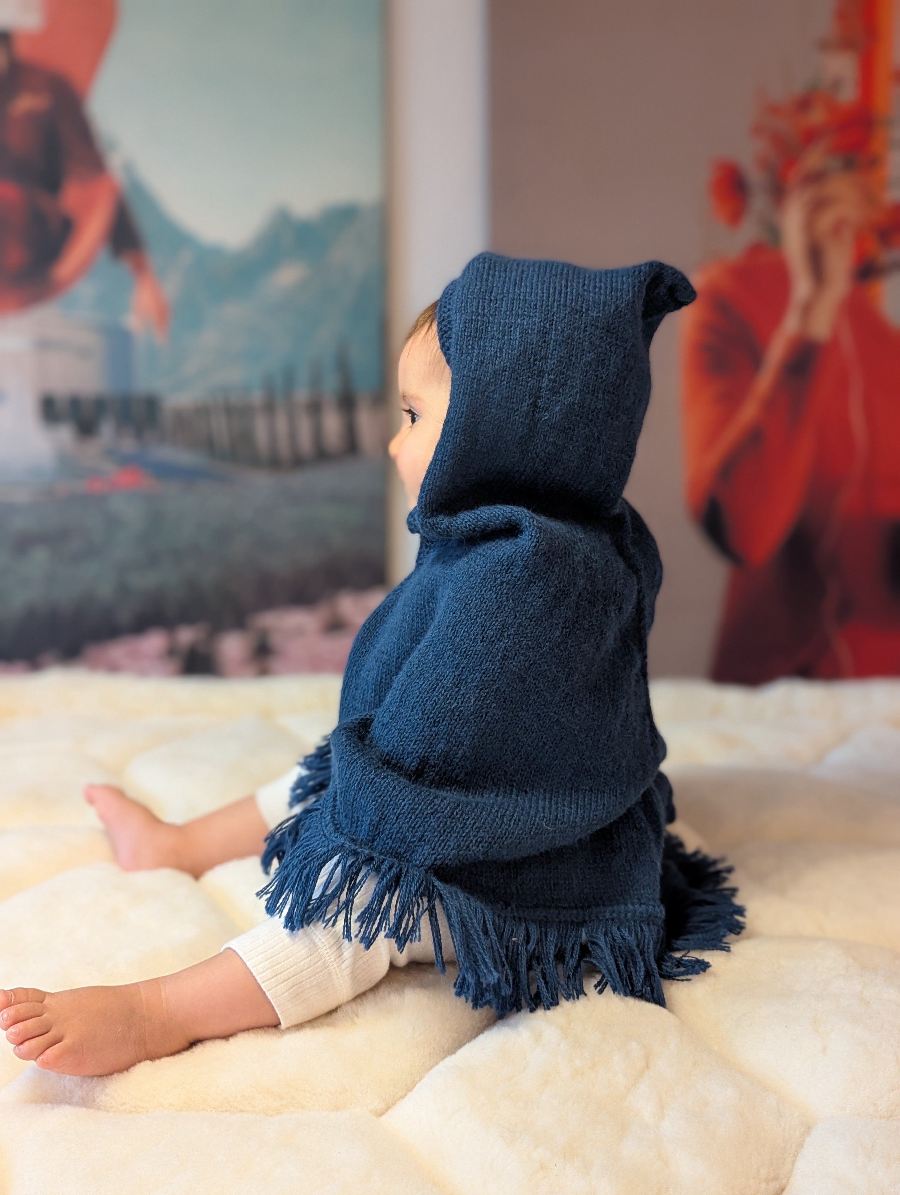 Child wearing a blue hooded poncho sitting on a soft surface with a colorful abstract painting in the background.