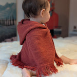 Child wearing a red hooded poncho sitting on a white surface with a blurred background