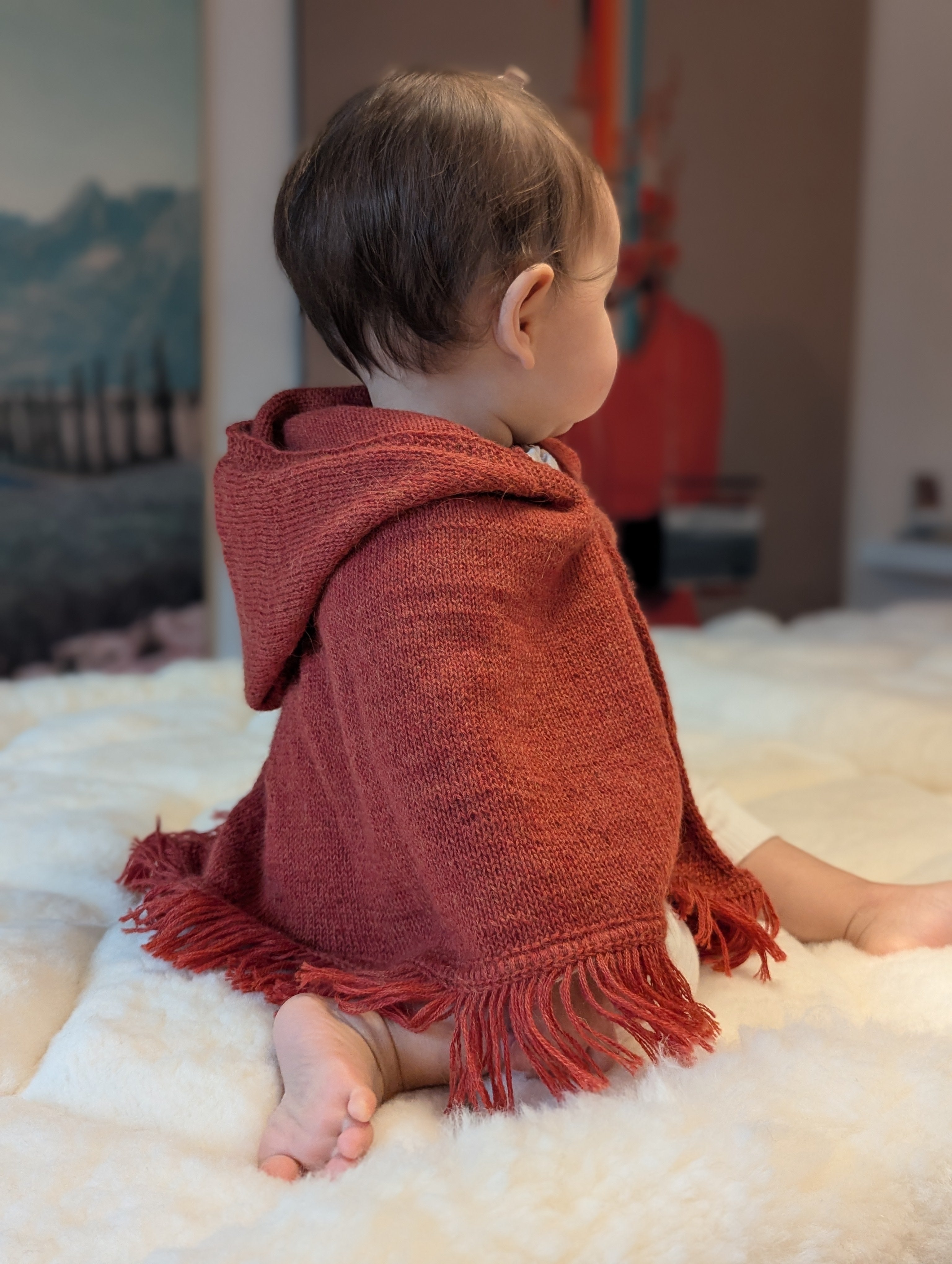 Child wearing a red hooded poncho sitting on a white surface with a blurred background