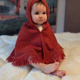 Baby wrapped in a red hooded blanket sitting on a white surface with a colorful painting in the background.