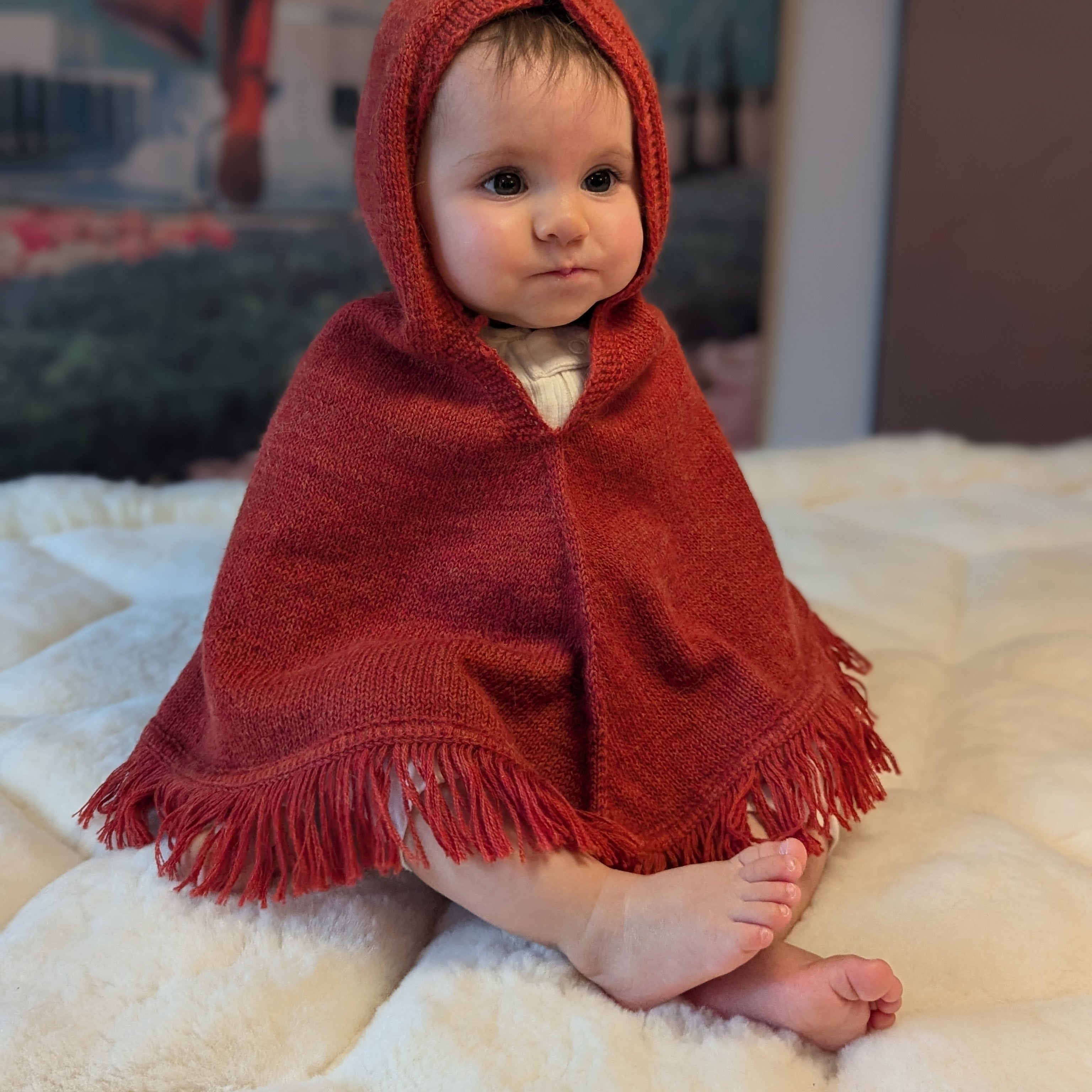 Baby wrapped in a red hooded blanket sitting on a white surface with a colorful painting in the background.