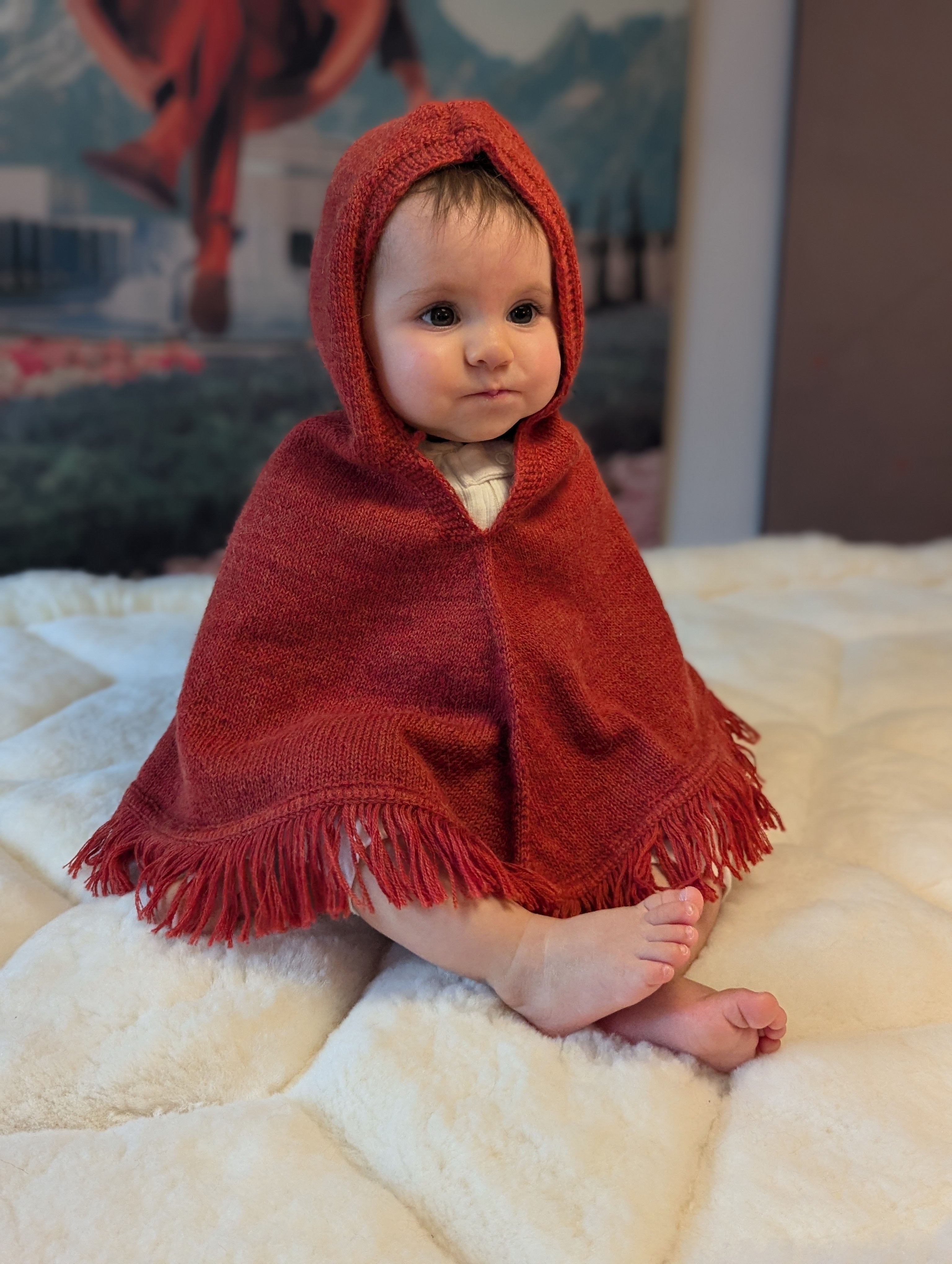 Baby wrapped in a red hooded blanket sitting on a white surface with a colorful painting in the background.