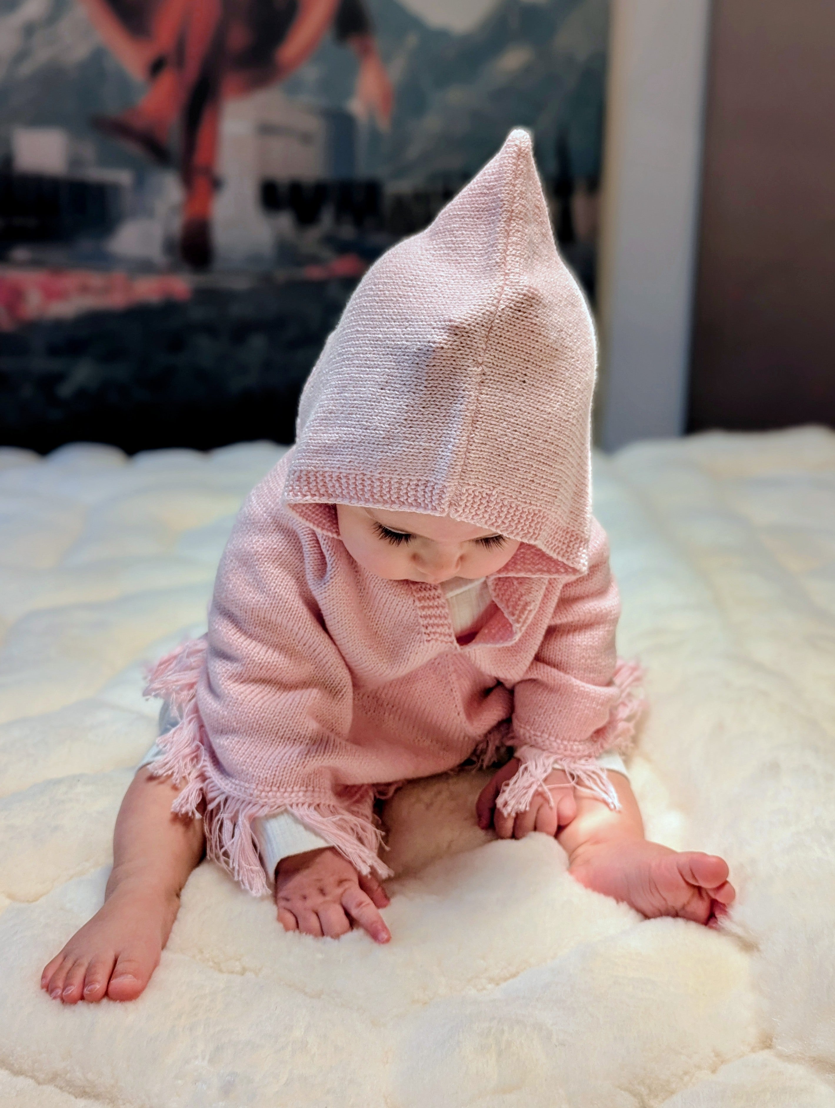 Baby wearing a pink knitted outfit with a hood on a white blanket.