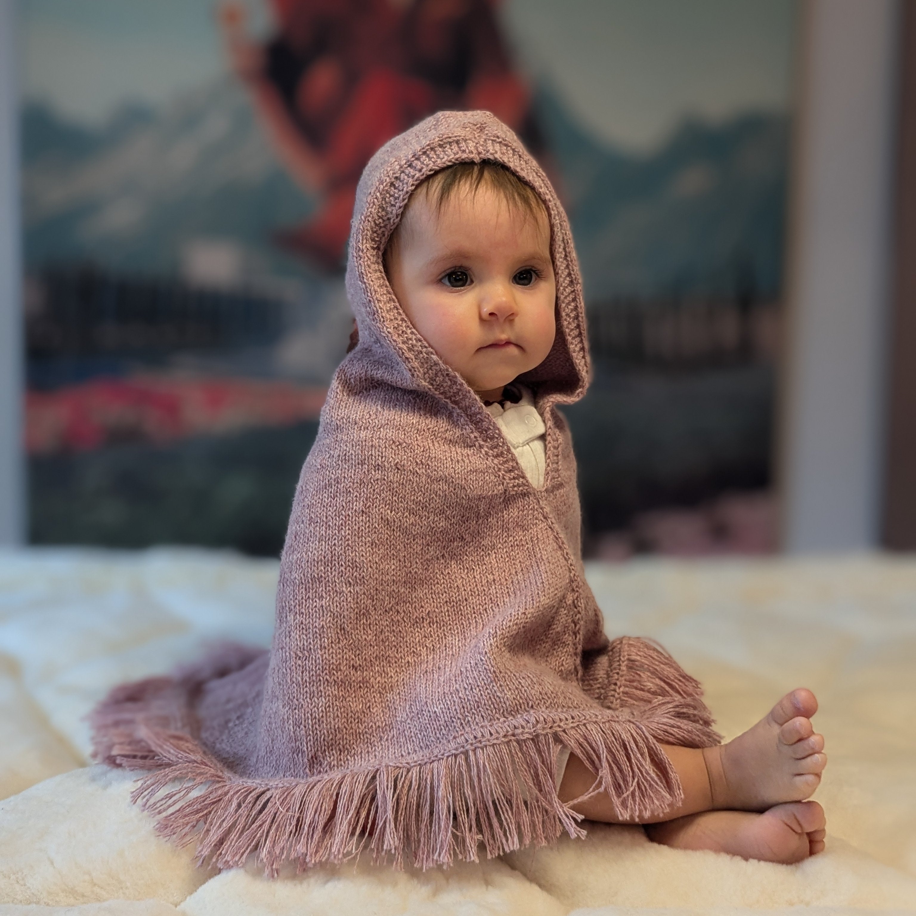 Baby wrapped in a pink hooded blanket sitting on a bed with a colorful painting in the background