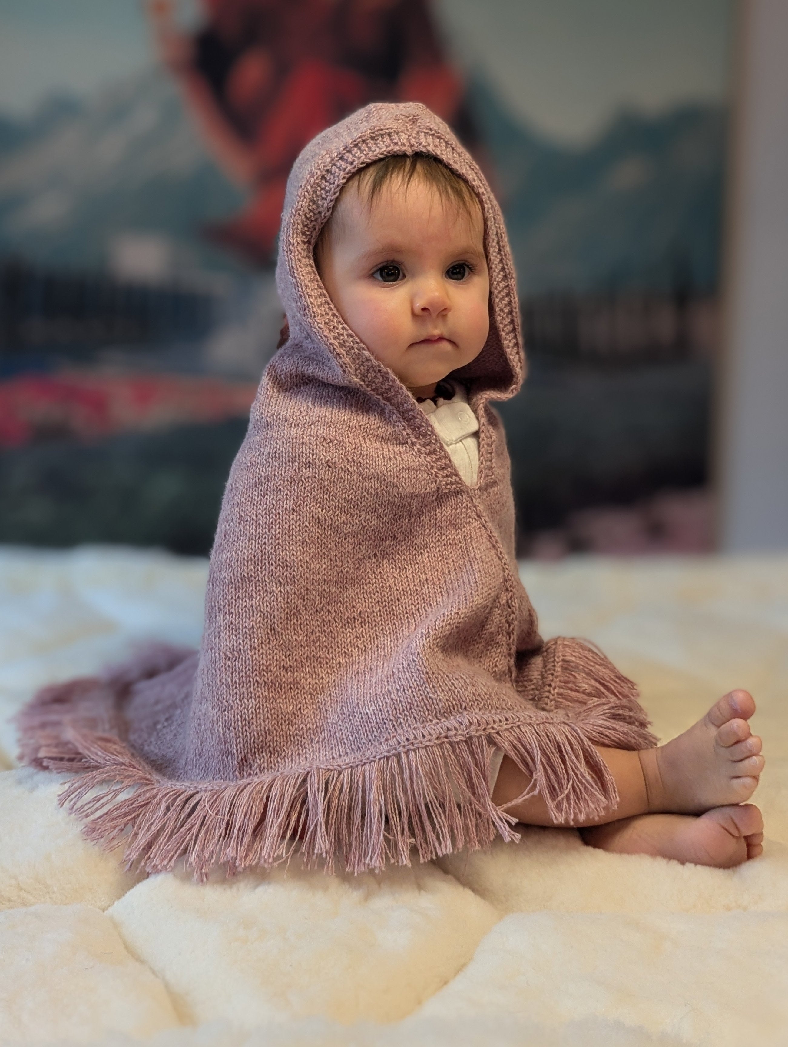 Baby wrapped in a pink hooded blanket sitting on a bed with a colorful painting in the background