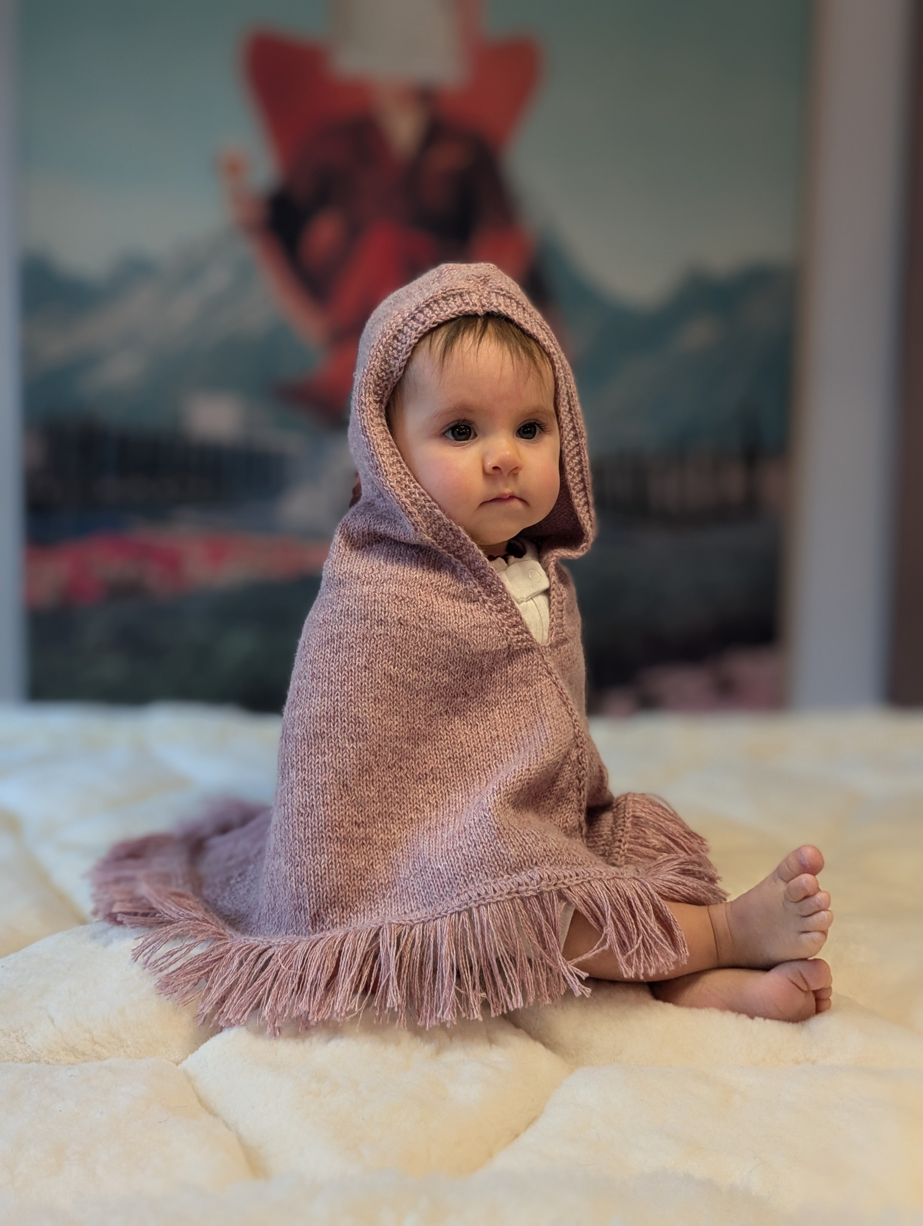Baby wrapped in a pink hooded blanket sitting on a bed with a colorful painting in the background