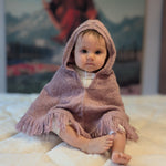 Baby in a pink hooded blanket sitting on a soft surface with a blurred background