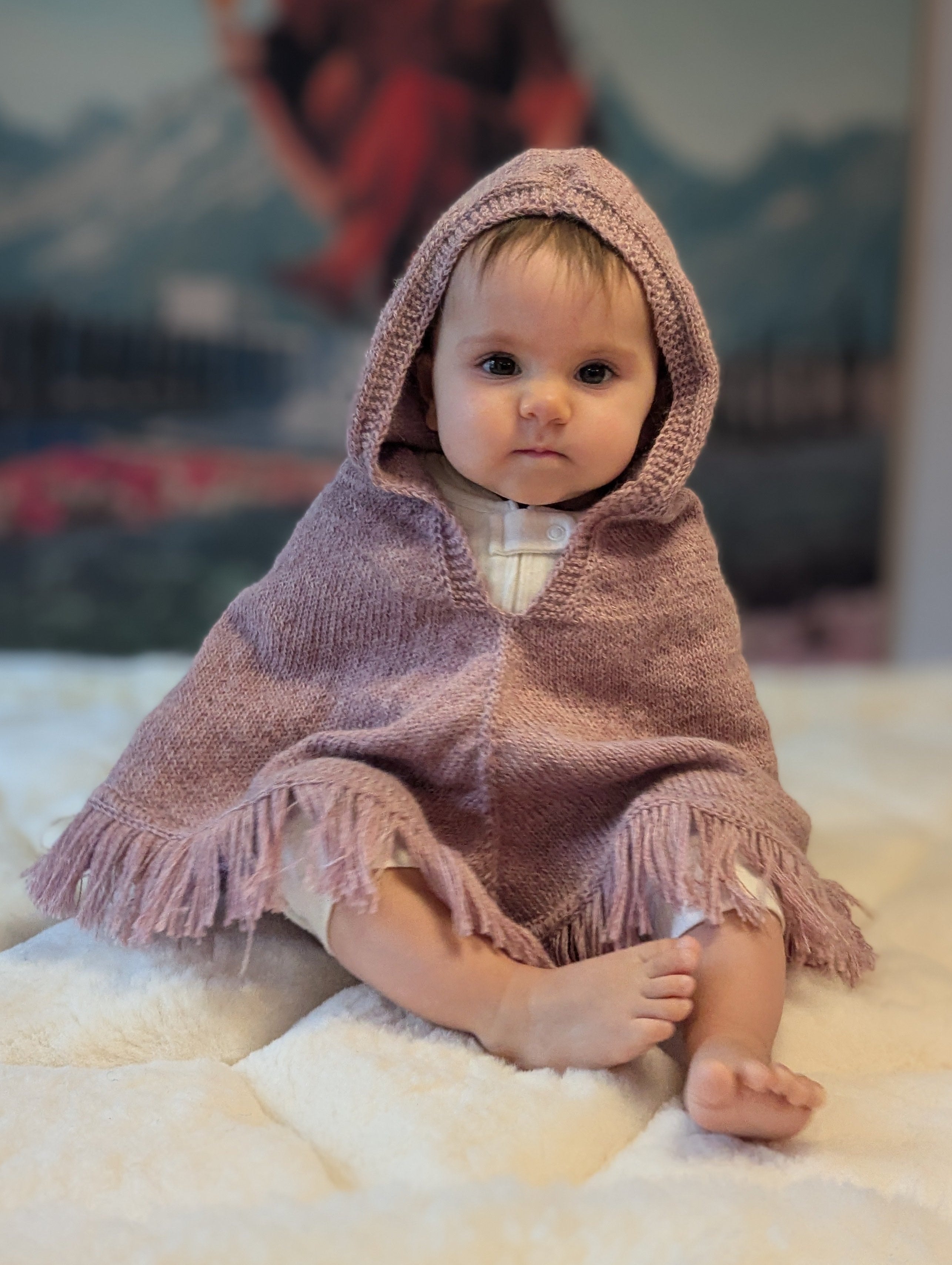 Baby in a pink hooded blanket sitting on a soft surface with a blurred background
