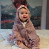 Baby wearing a pink hooded blanket sitting on a white surface with a colorful painting in the background.