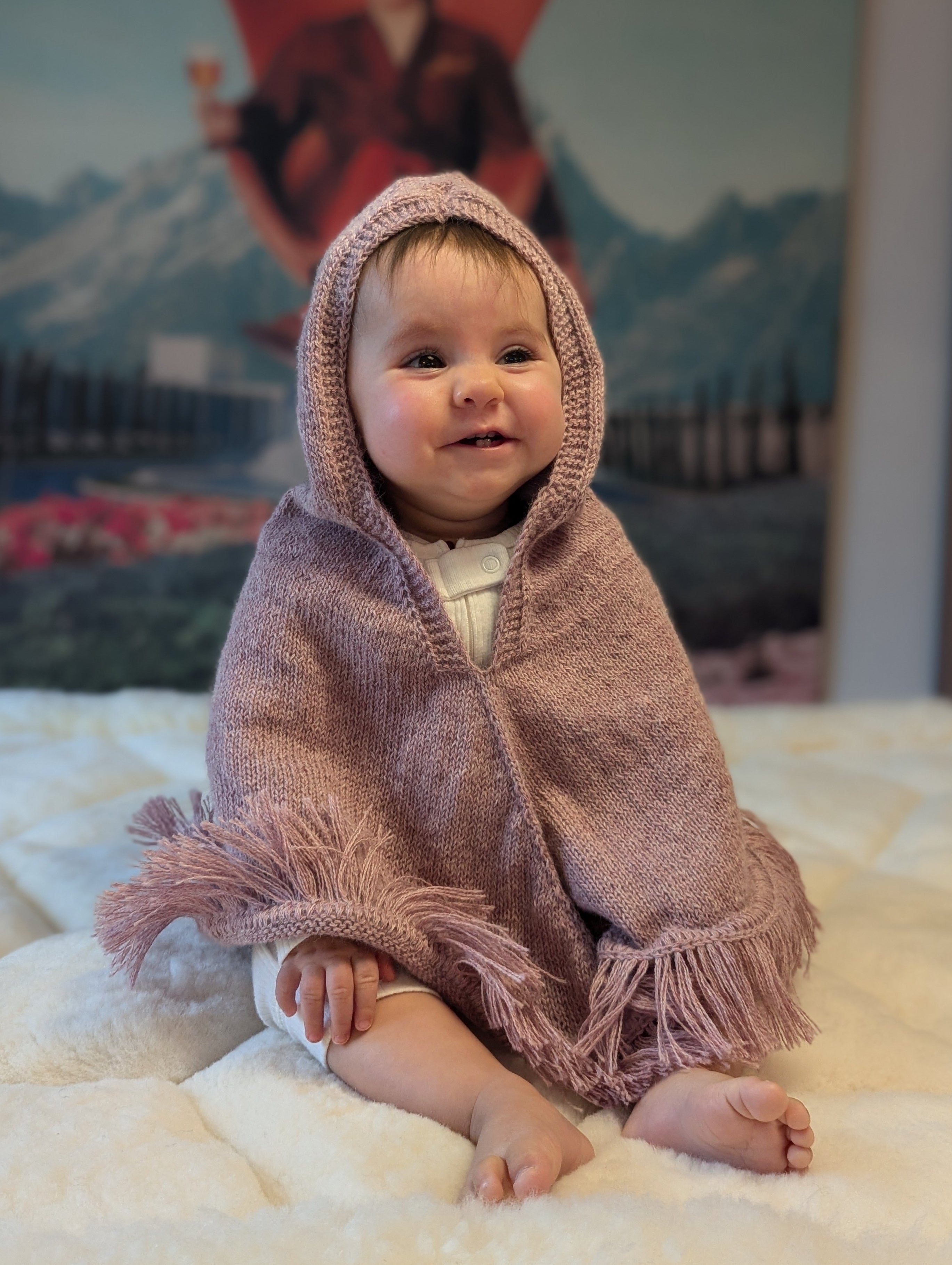Baby wearing a pink hooded blanket sitting on a white surface with a colorful painting in the background.
