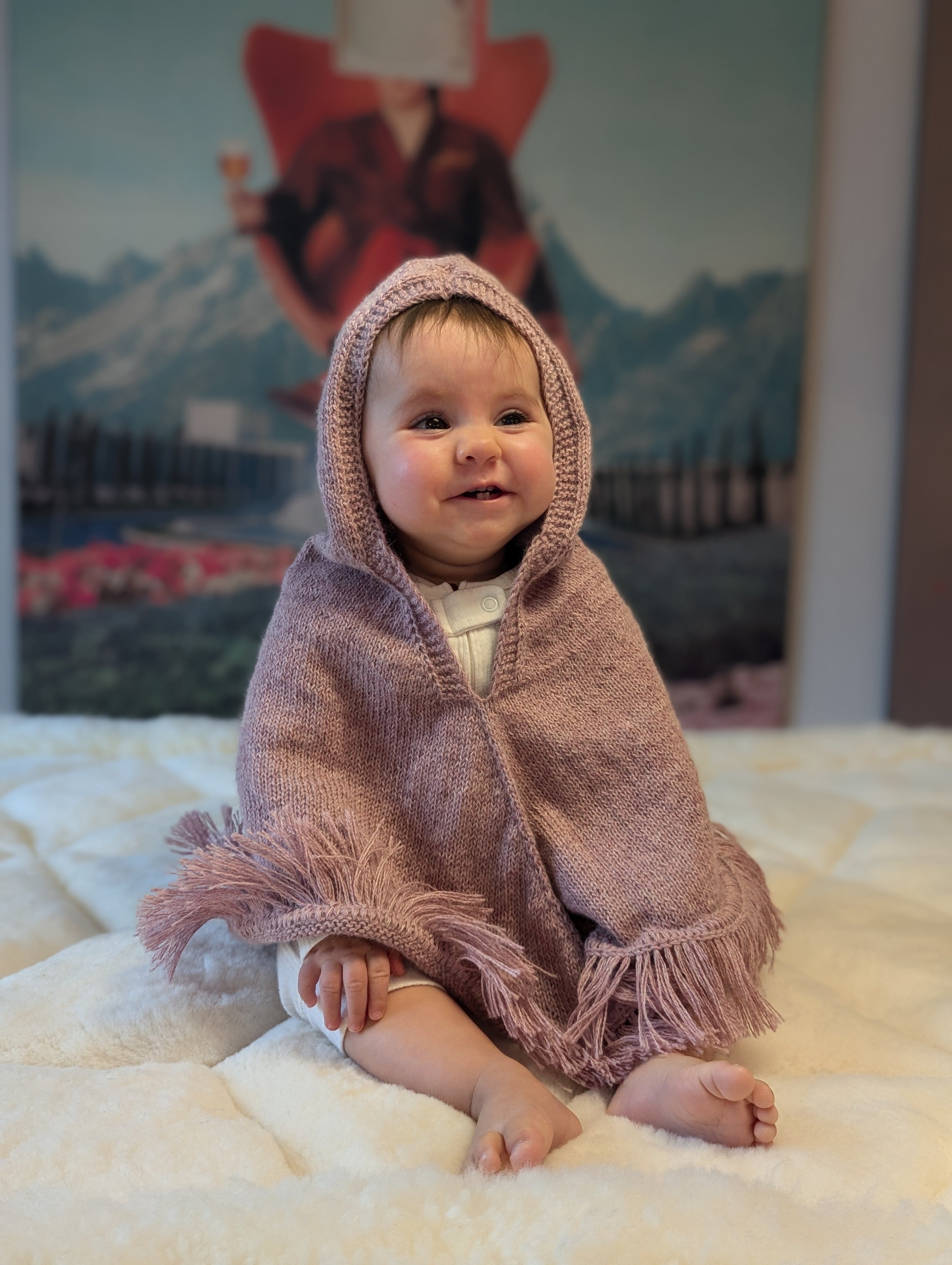 Baby wearing a pink hooded blanket sitting on a white surface with a colorful painting in the background.