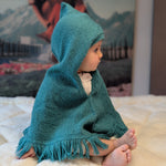 Child wearing a teal hooded poncho sitting on a bed with a mountain-themed wallpaper in the background.