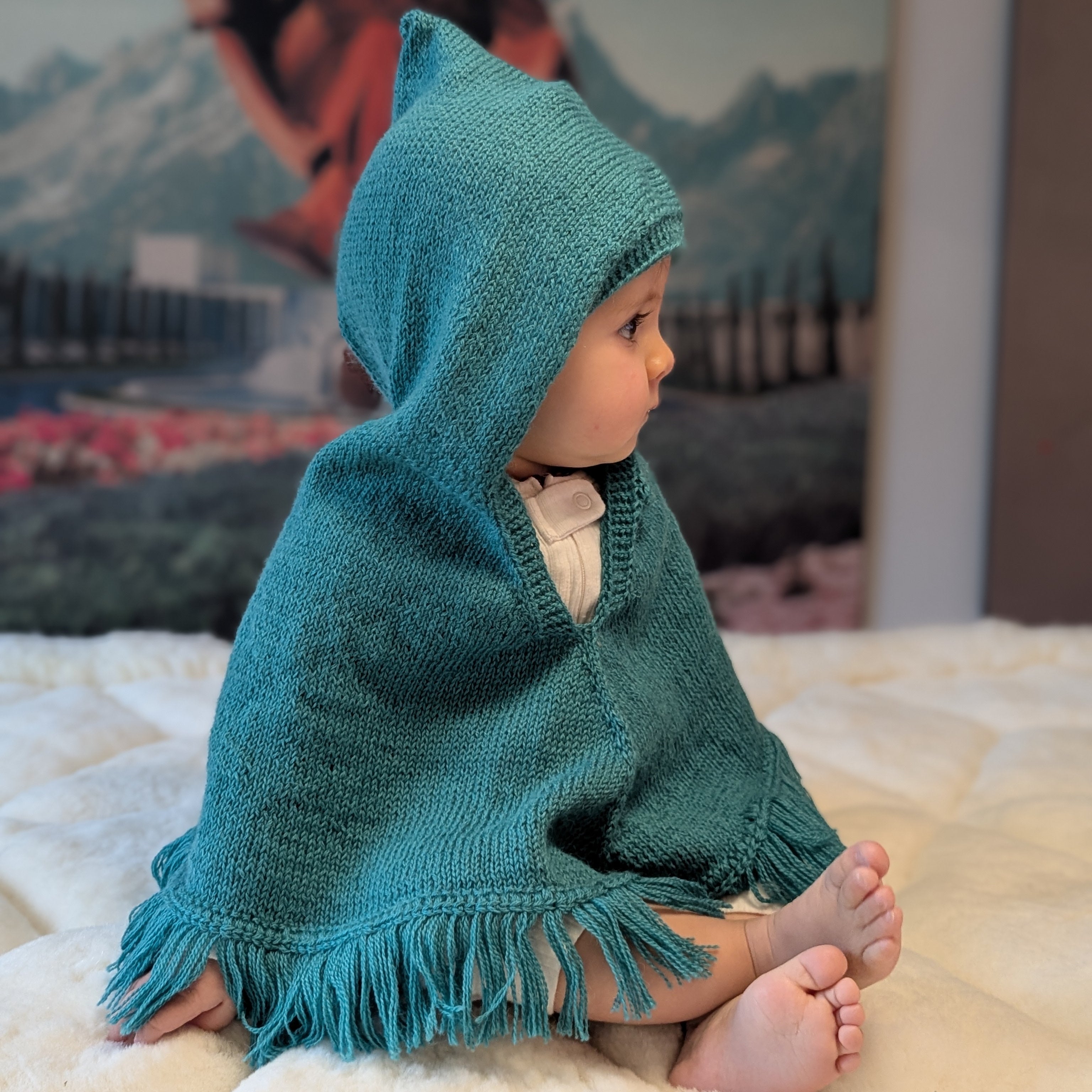 Child wearing a teal hooded poncho sitting on a bed with a mountain-themed wallpaper in the background.