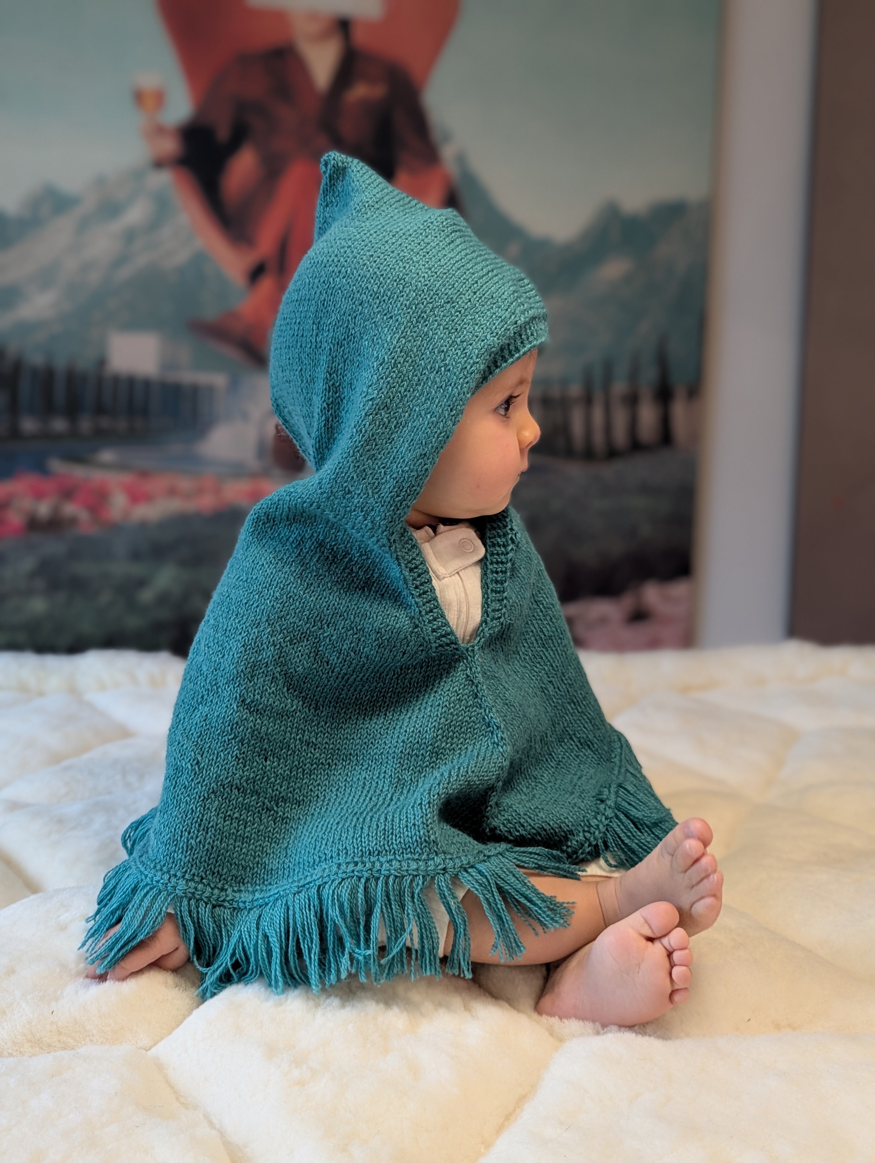 Child wearing a teal hooded poncho sitting on a bed with a mountain-themed wallpaper in the background.