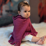 Baby sitting on a white blanket wearing a purple poncho with tassels.