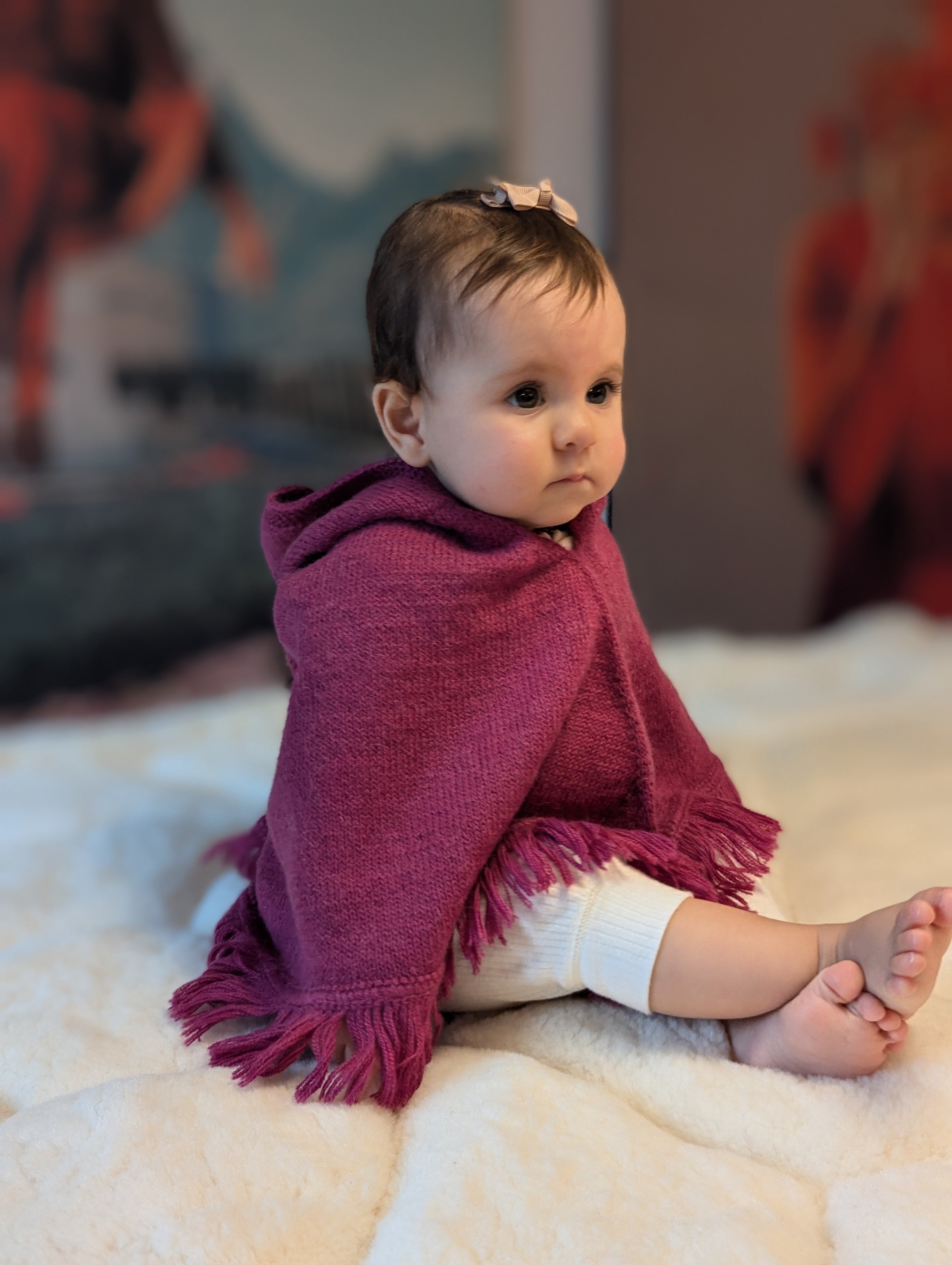 Baby sitting on a white blanket wearing a purple poncho with tassels.
