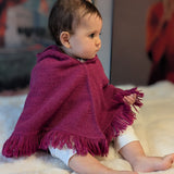 Baby wearing a purple poncho with fringe sitting on a white surface.